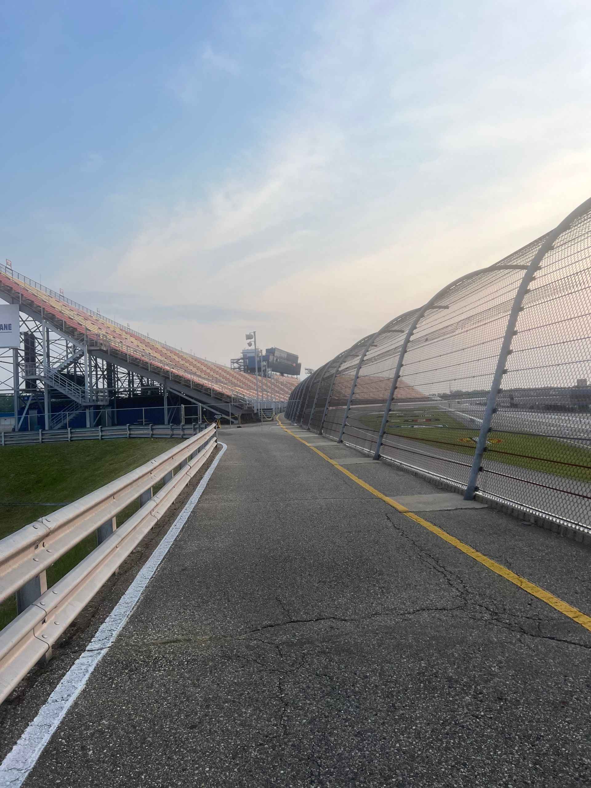 Asphalt path between race track grandstand and safety netting. Cloudy blue sky overhead.