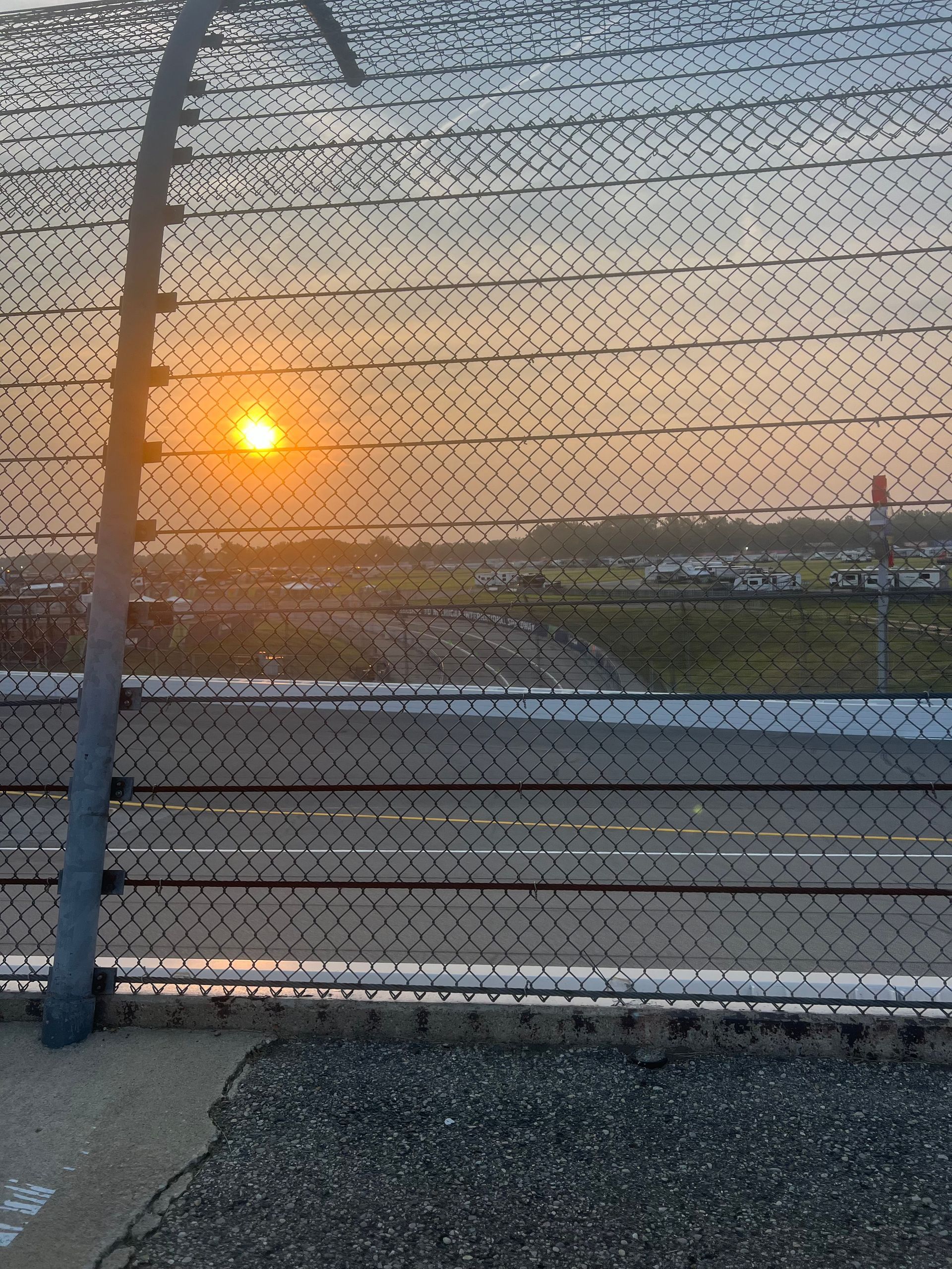 Sunset view through a chain link fence. Bright sun over a flat, open area, possibly an airport.