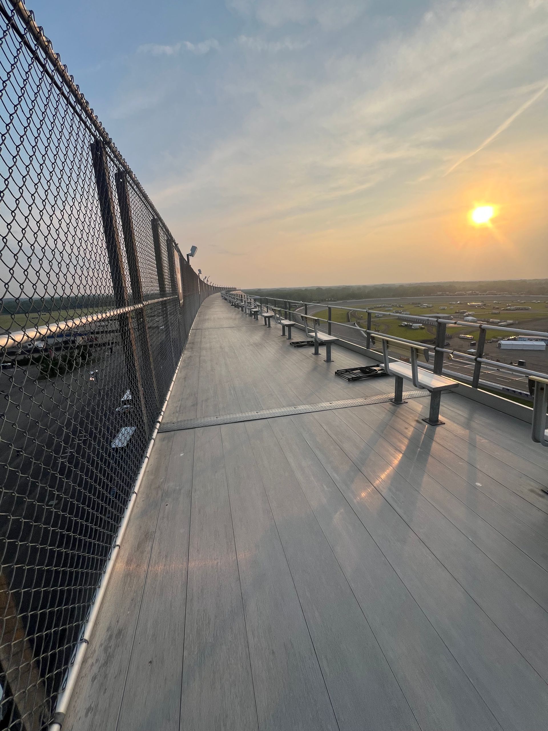 View from a high walkway with metal grating, chain-link fence, and the sun setting.