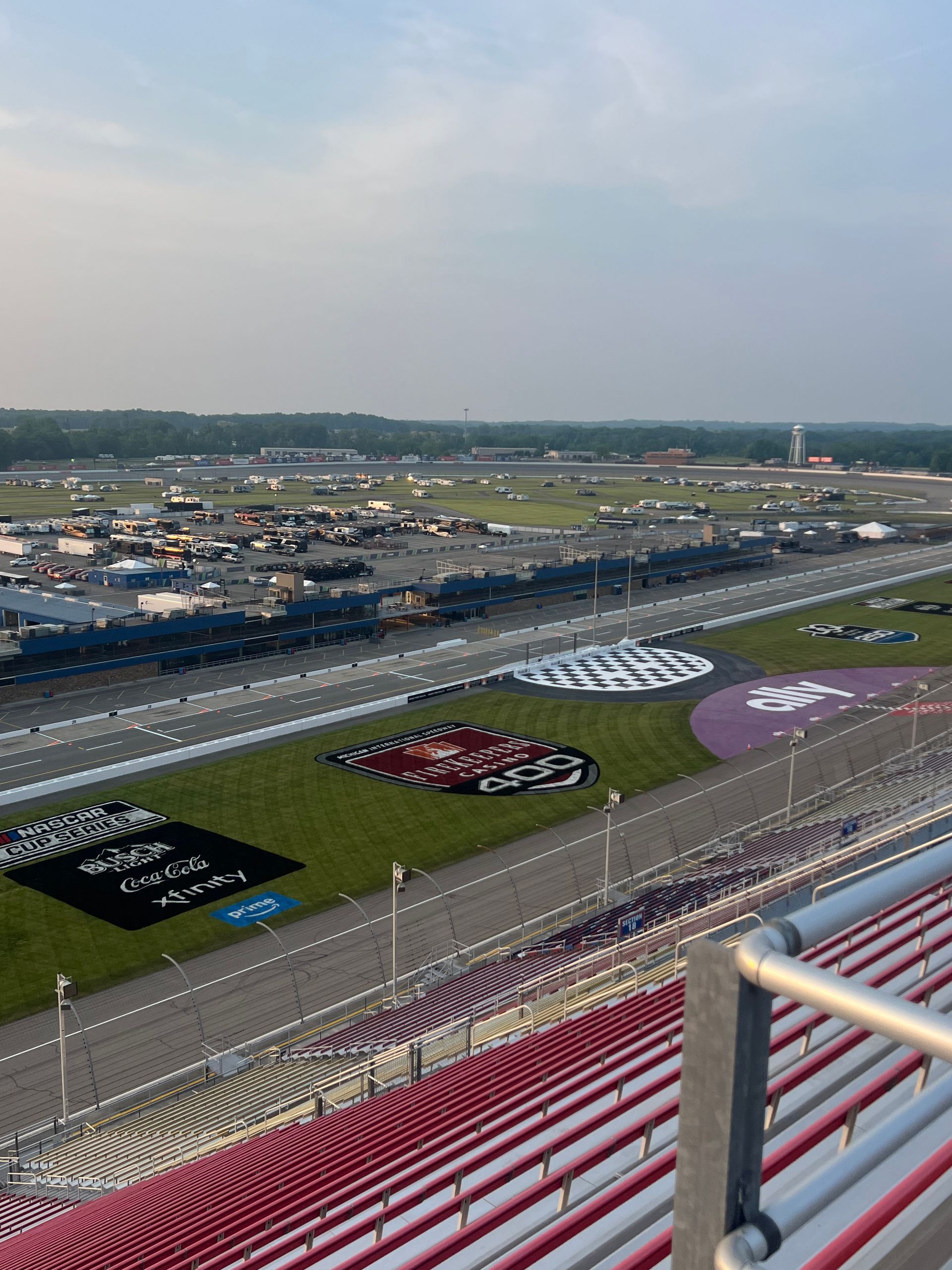 View of a racetrack with green infield, stands, and various vehicles; overcast sky.