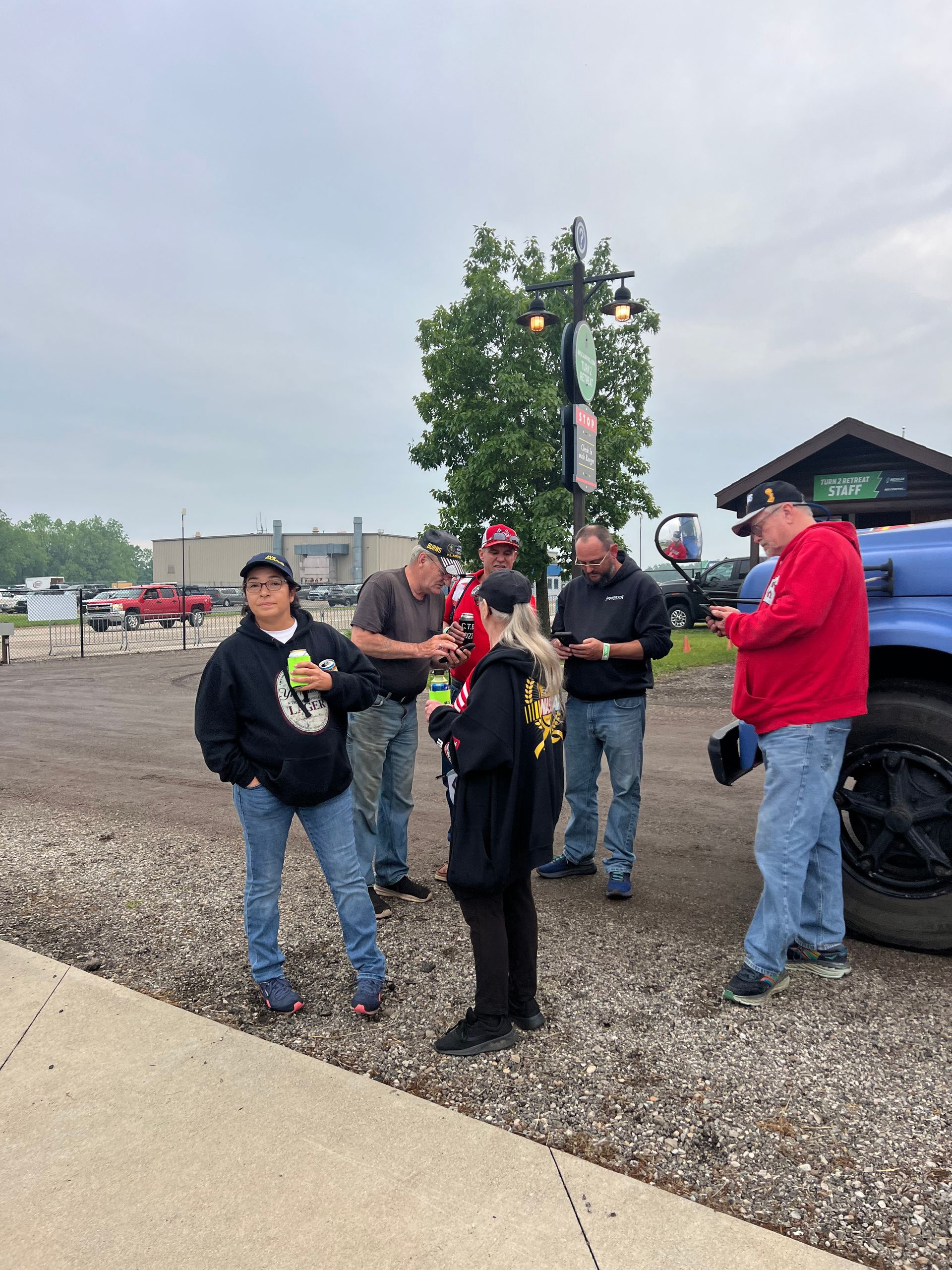 Group of people outside a business. Some hold drinks, look at phones, near a large truck and a green tree.