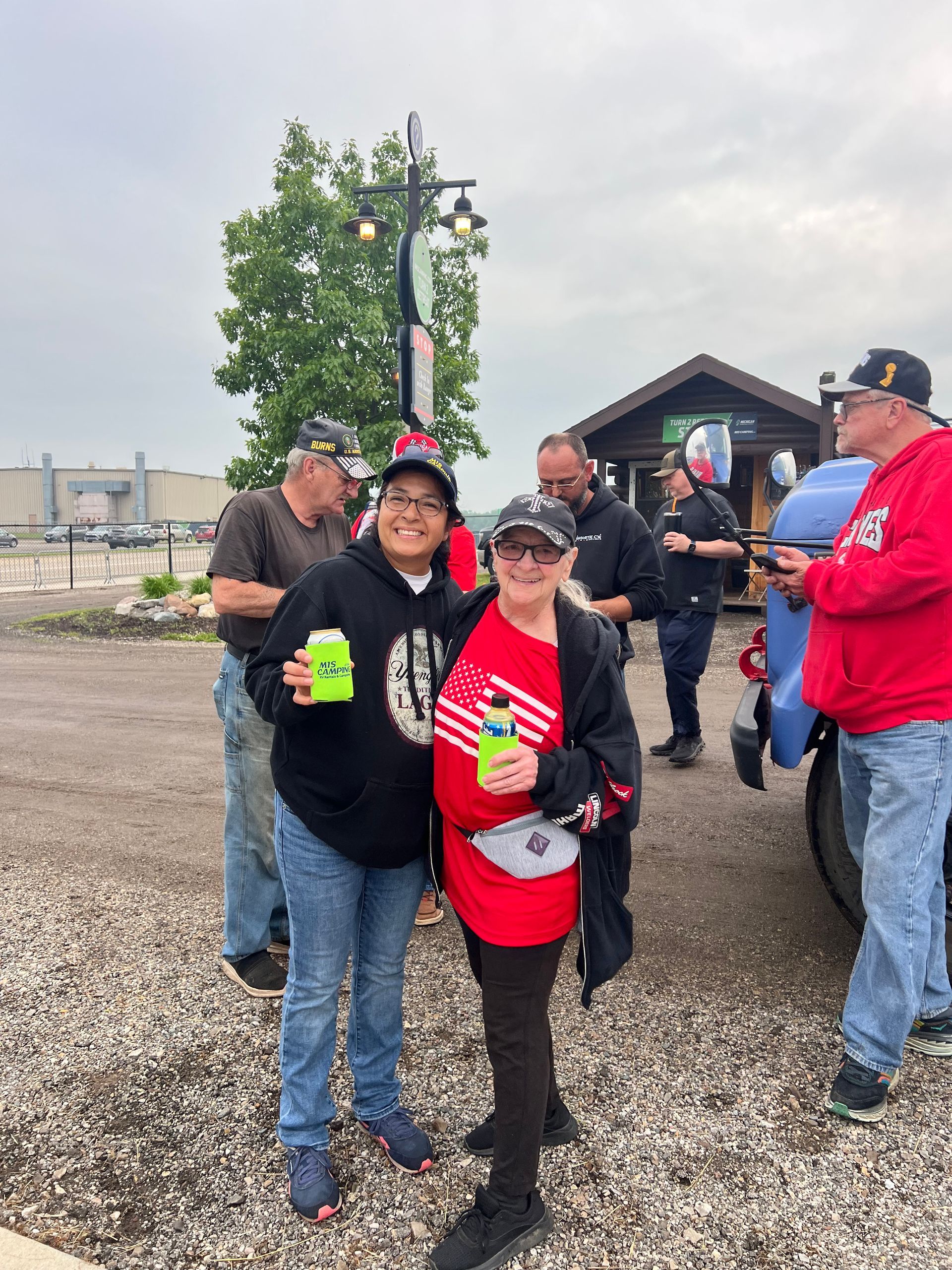 Two people holding cans, smiling, pose outside with others near a building and blue truck. Cloudy day.