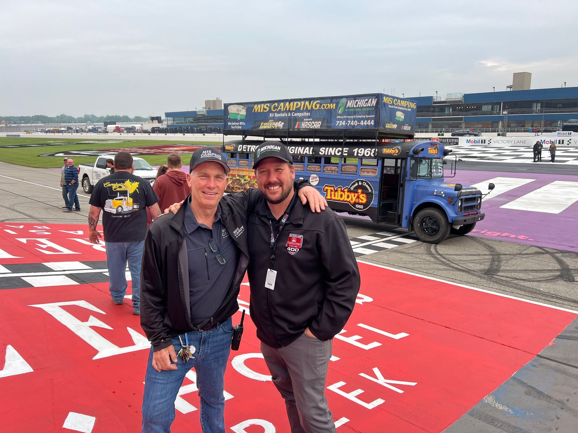 Two men pose in front of a blue school bus decorated with signage; racetrack in the background.