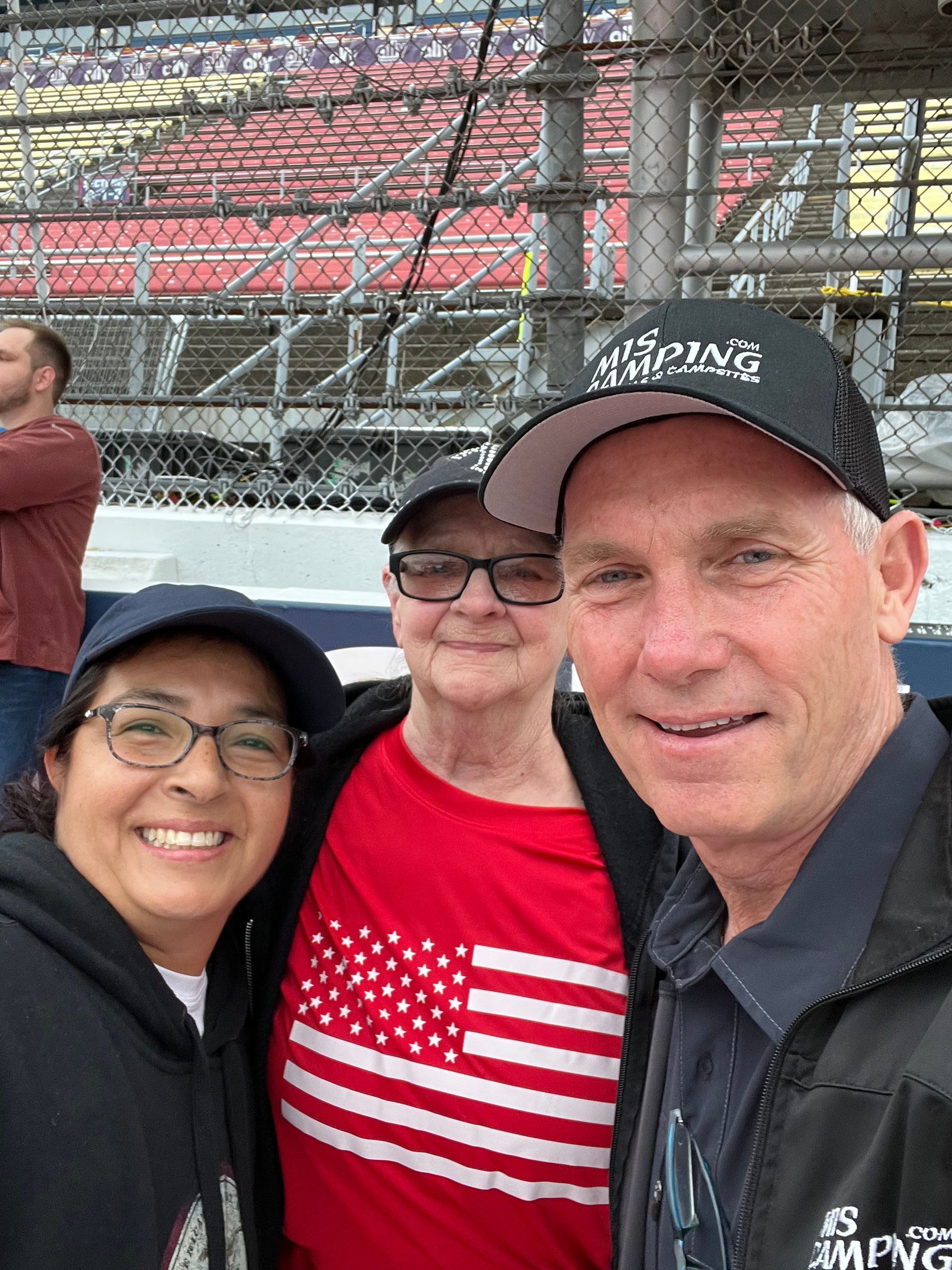Three people smiling at a racetrack. One wears an American flag shirt. They're near a chain-link fence.