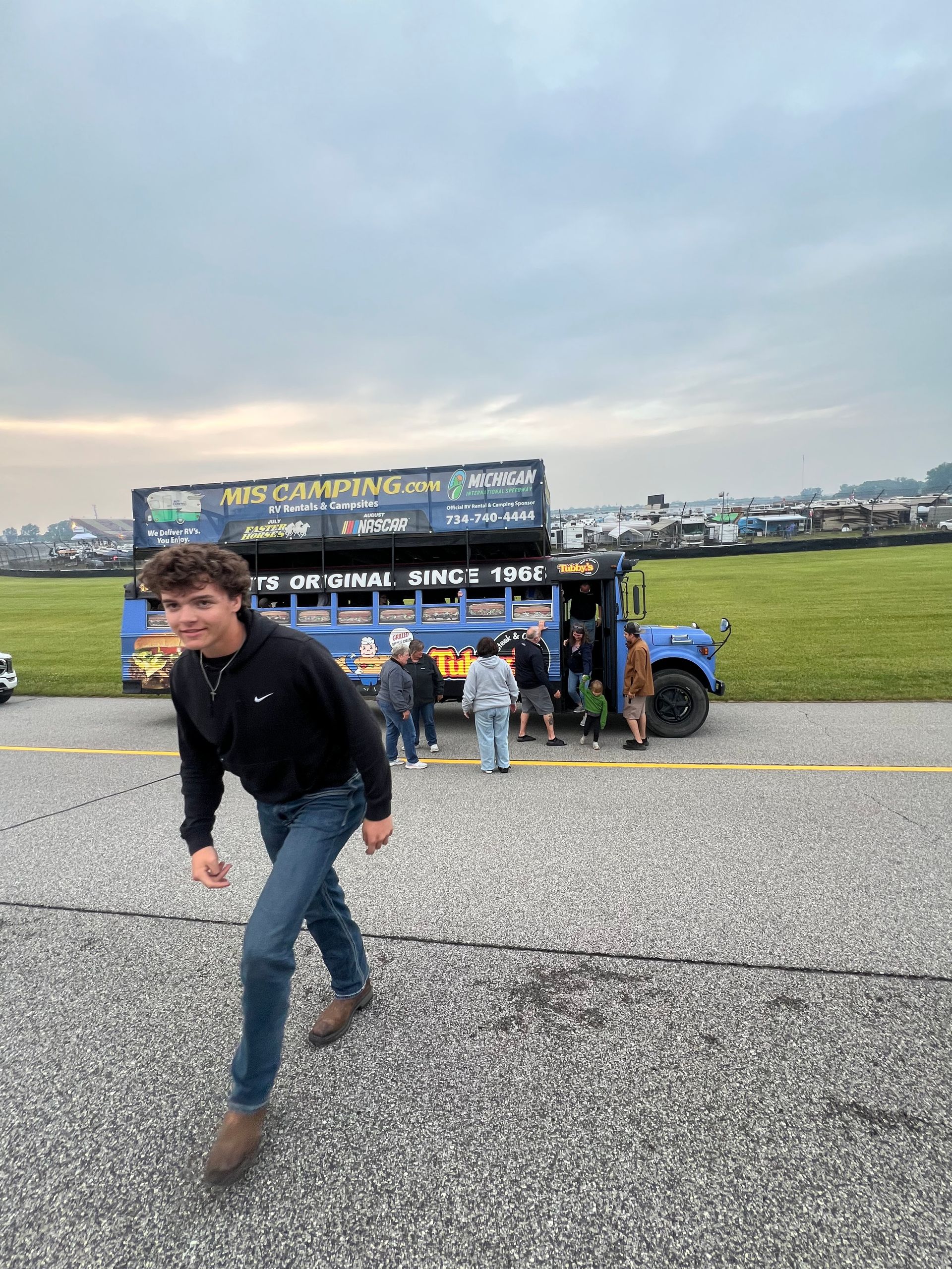 Man walking towards blue food truck, people waiting. Asphalt, grass, overcast sky.