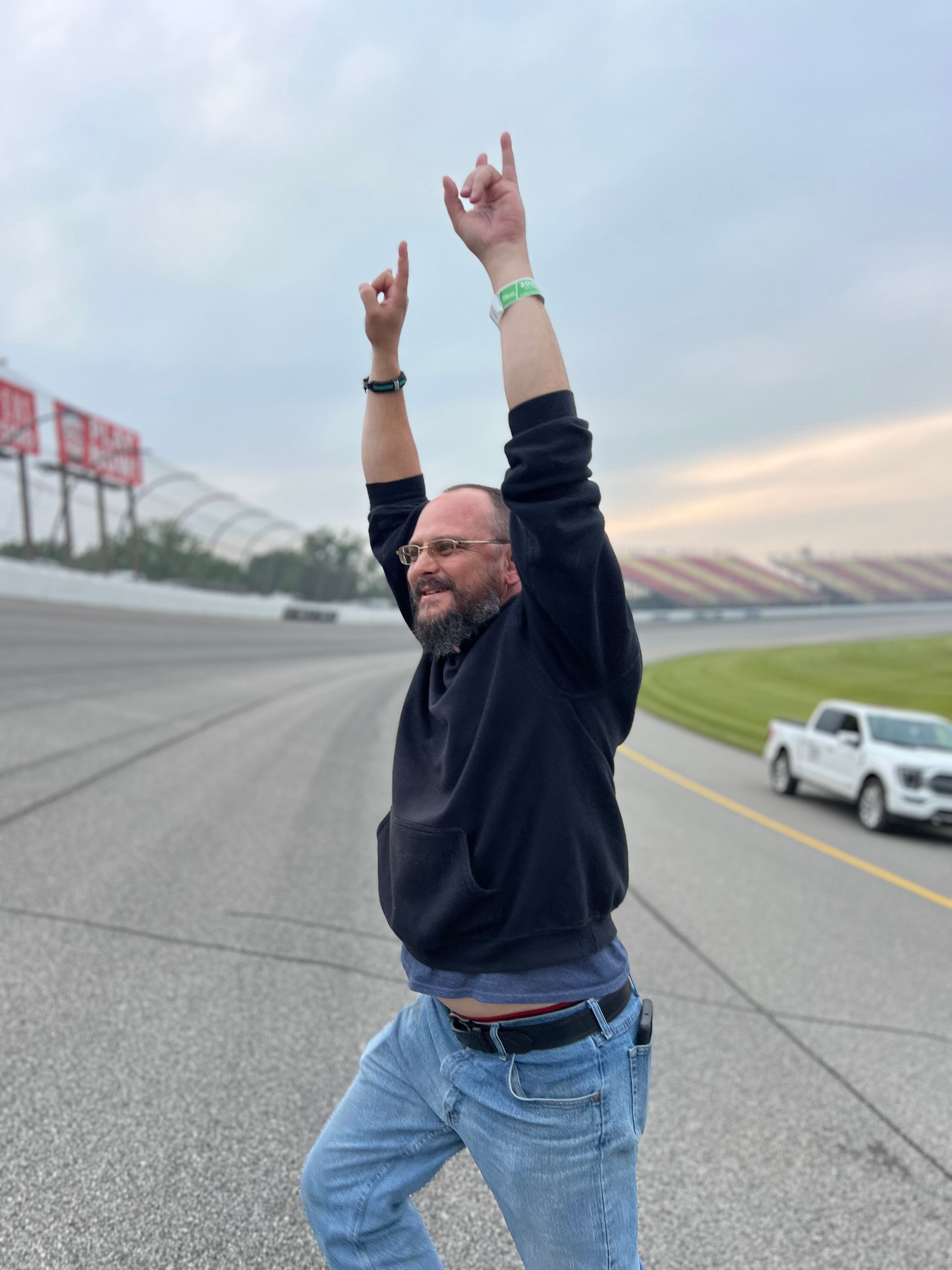 Man with arms raised, standing on a racetrack. He wears a black hoodie, blue jeans, and a wristband. Cloudy sky.