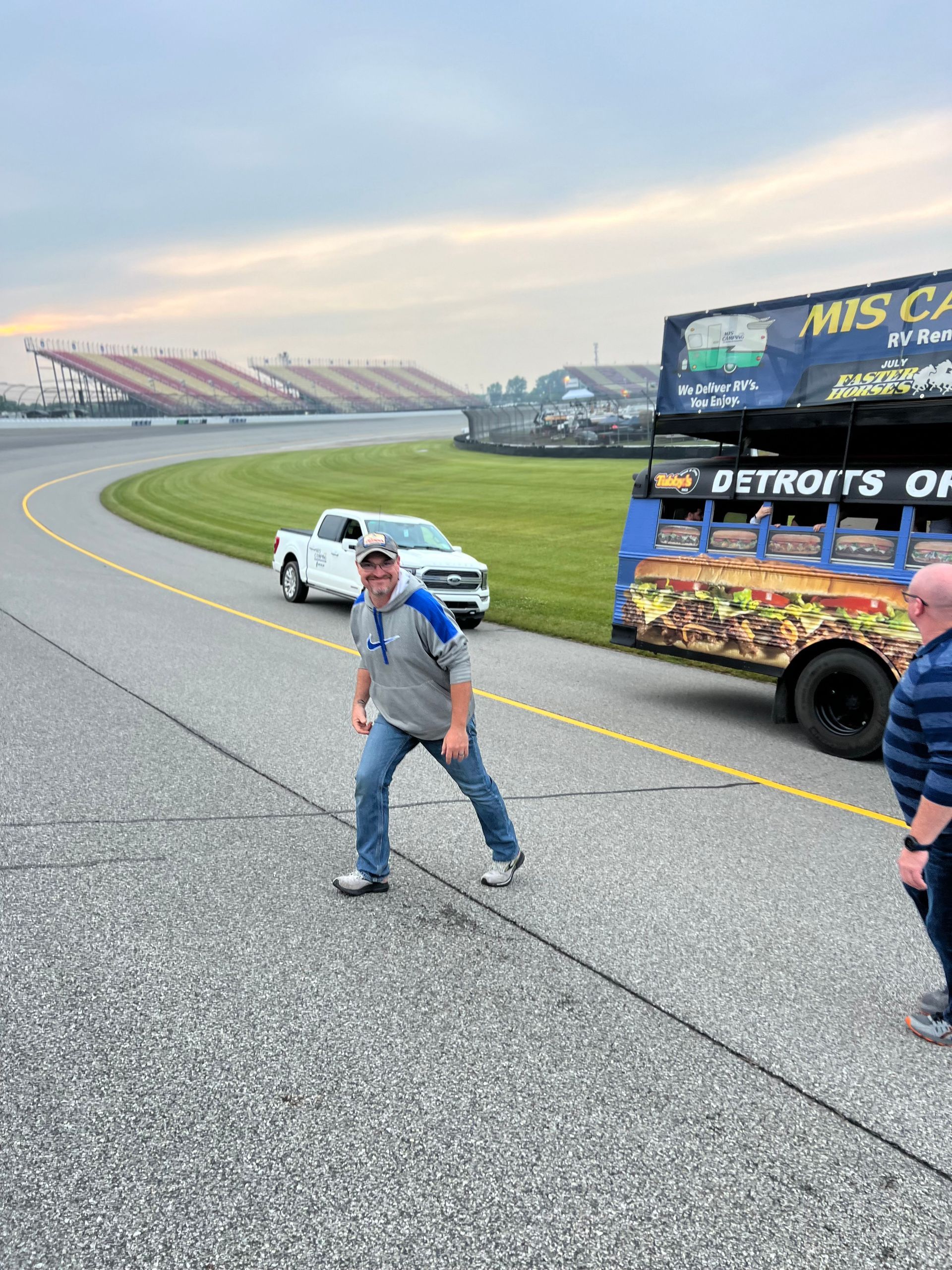Man on racetrack, Detroit bus nearby, white pickup truck. Cloudy sky, stadium in the background.