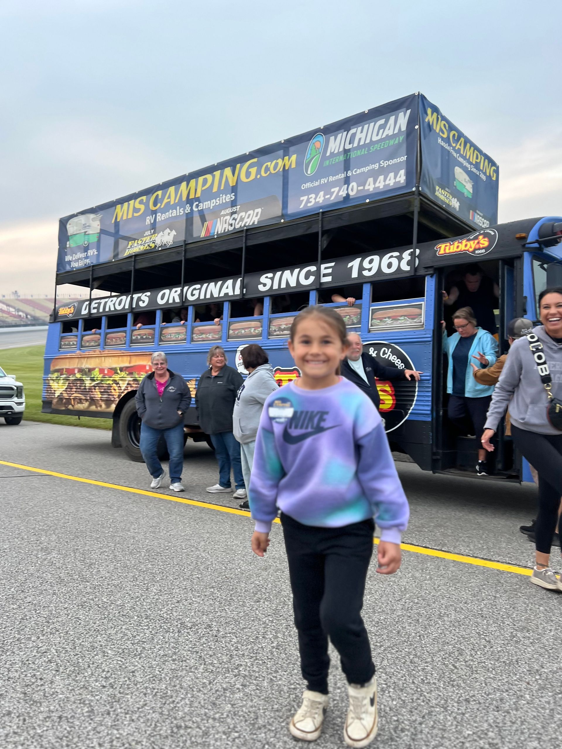 Girl in tie-dye sweatshirt smiles in front of a blue bus with 