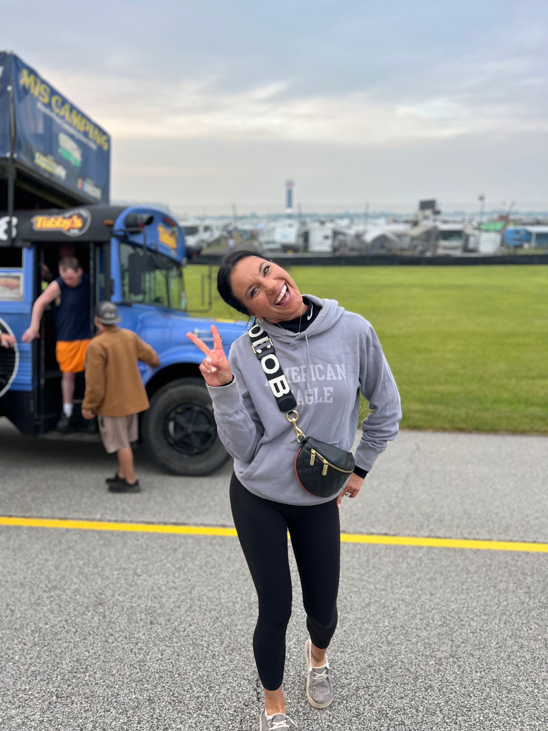Woman in gray hoodie makes peace sign, by a blue bus on an airfield.