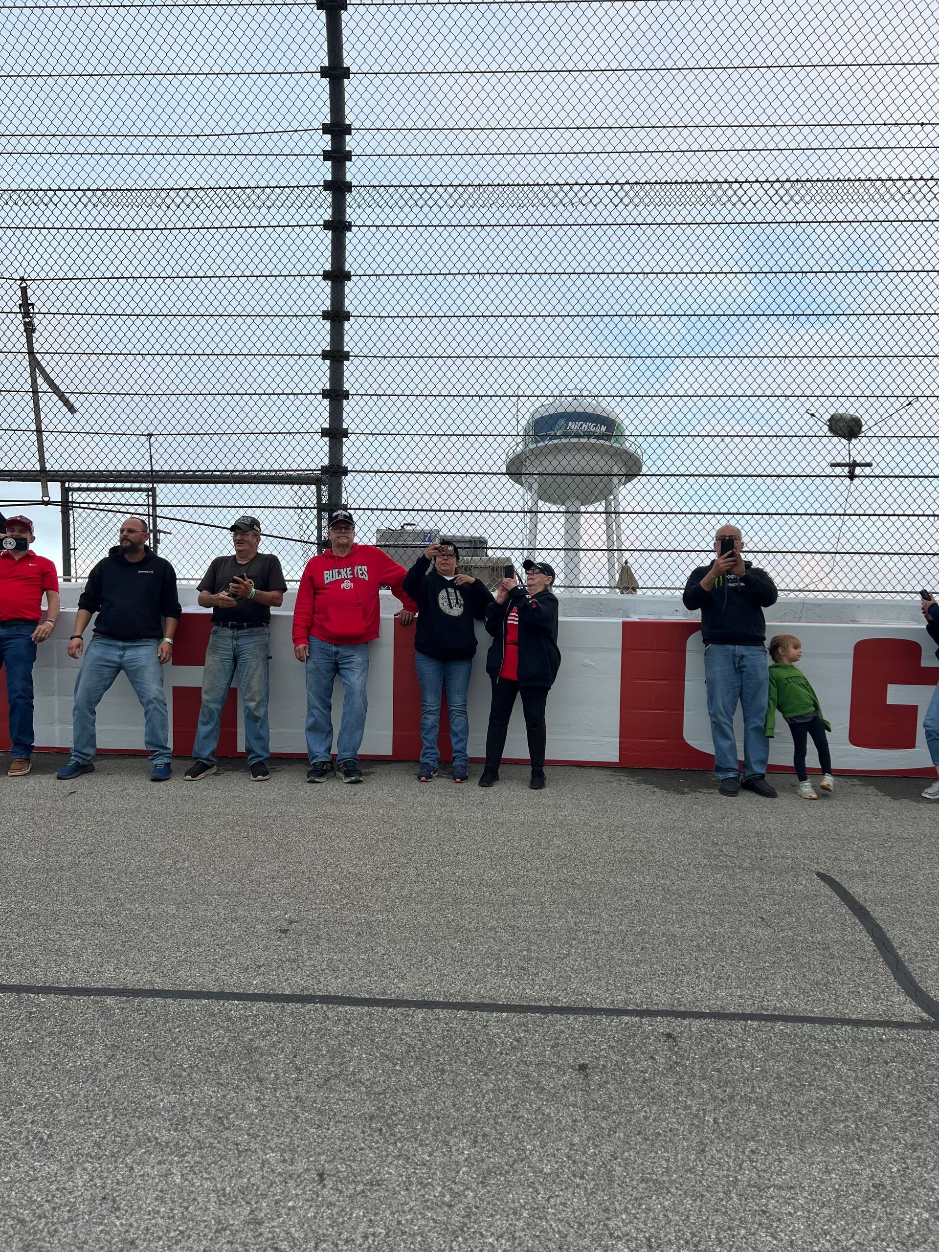 People standing in front of a racetrack fence. A control tower is visible in the background.