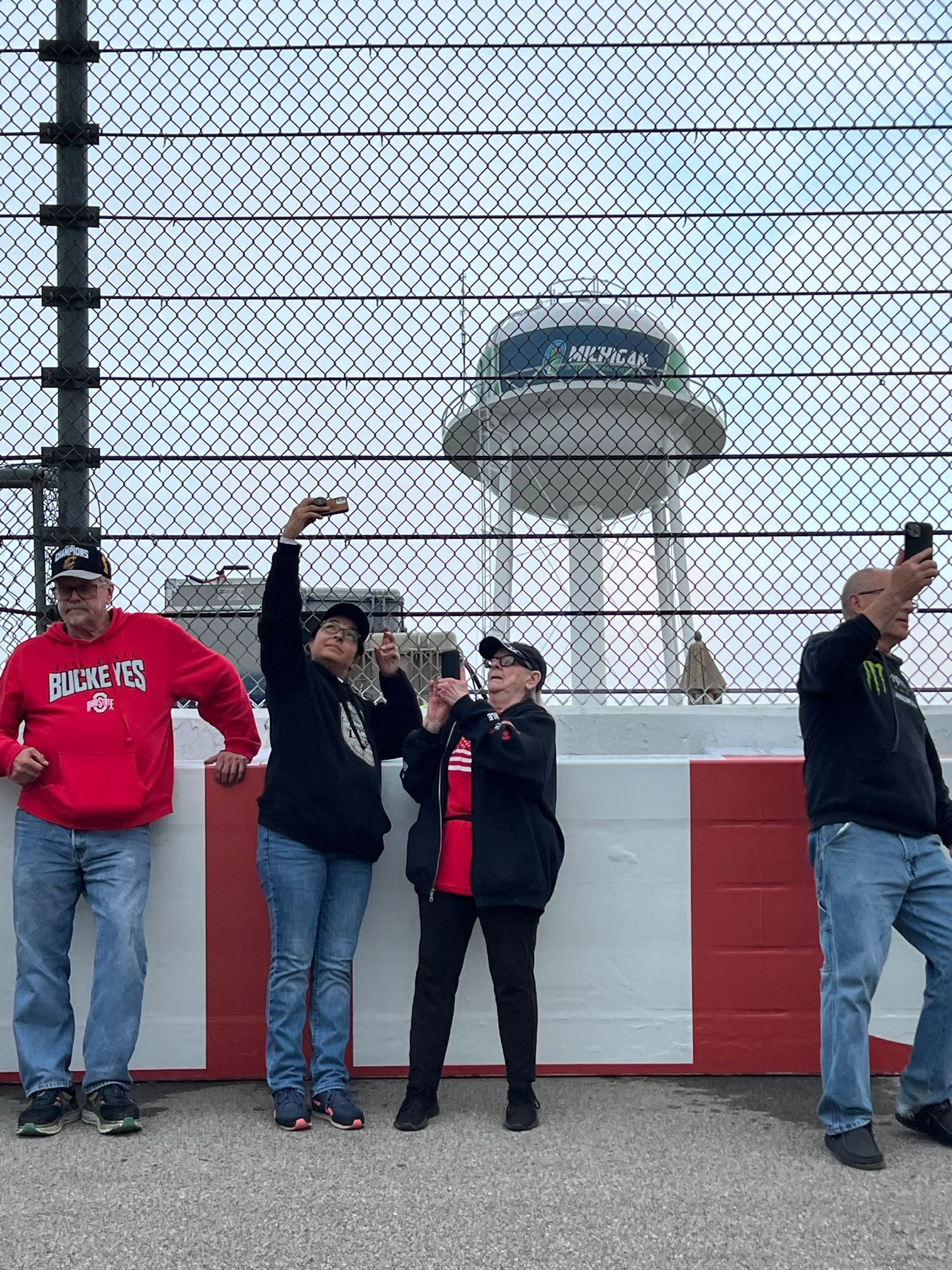 People stand near a chain-link fence, taking photos of a control tower at a racetrack.