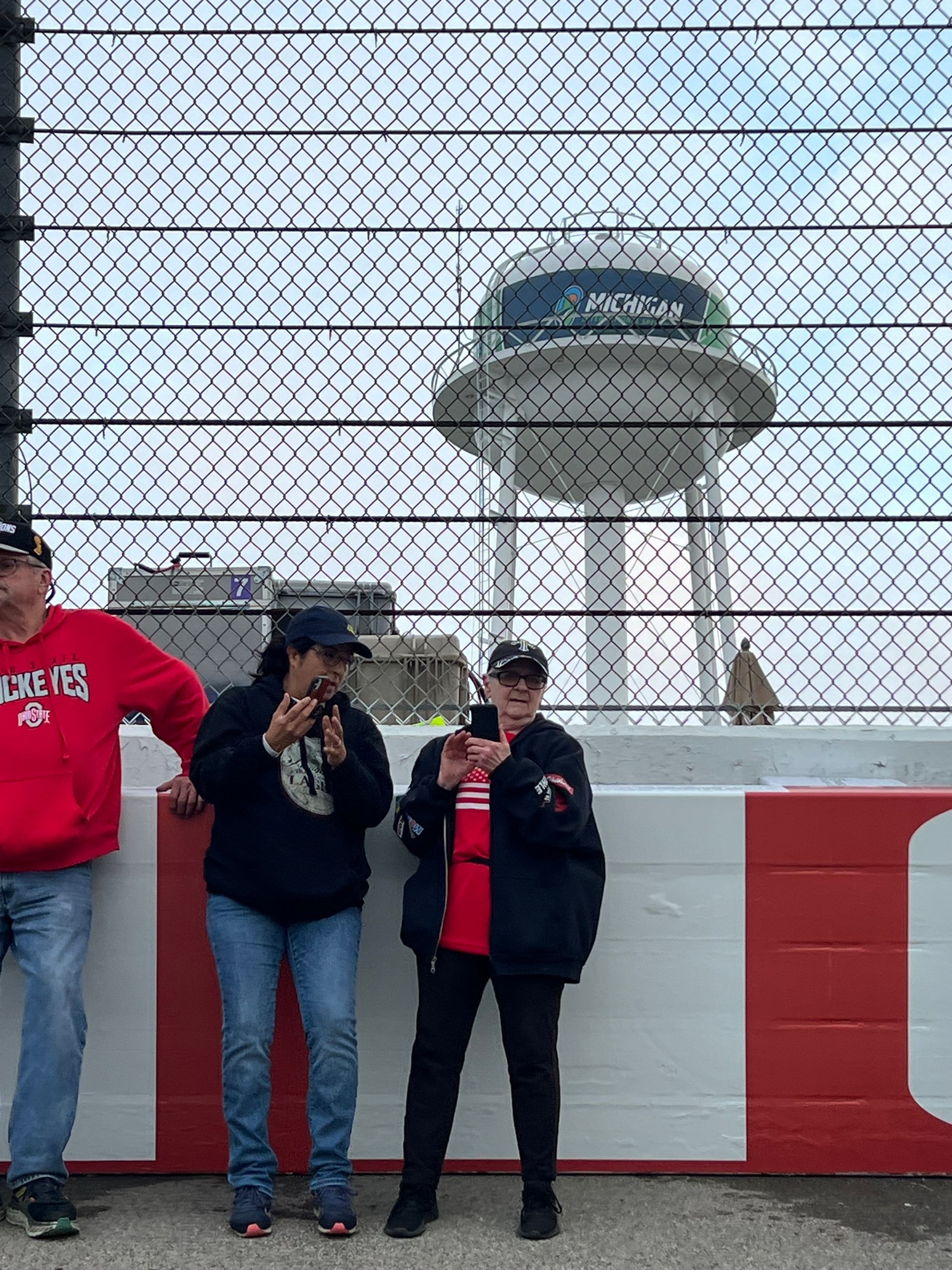 Three people stand behind a fence, looking at something off-camera. A white and green tower is in the background.