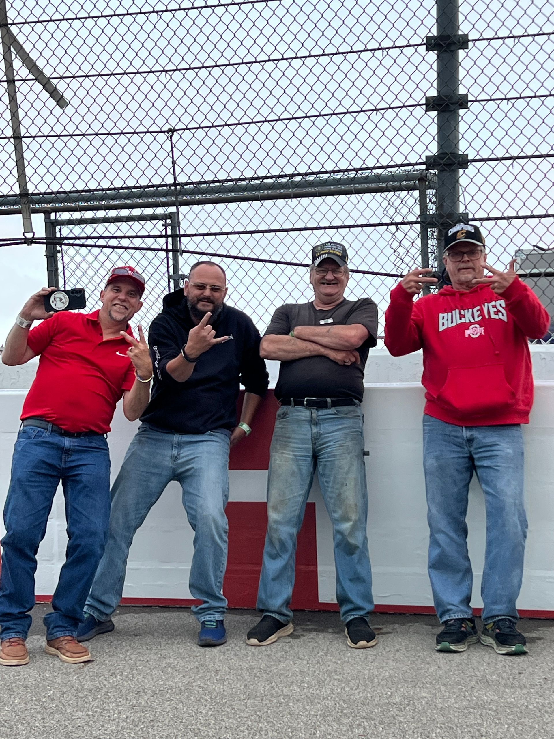 Four men posing by a fence and concrete barrier. They are making gestures, wearing casual clothes, and in an outdoor setting.