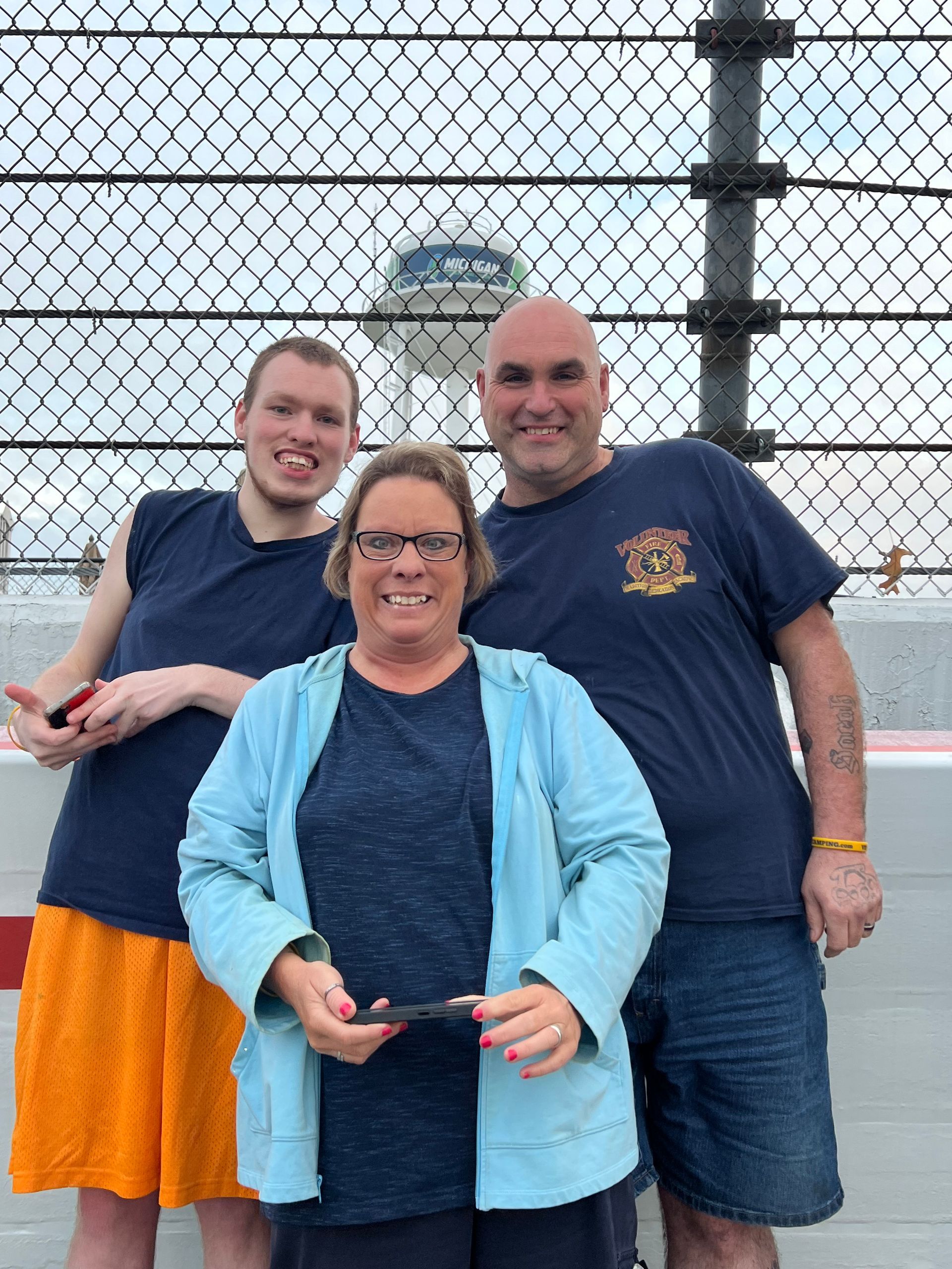 Three people posing in front of a chain-link fence and airport control tower. Woman in blue jacket smiles between two men.