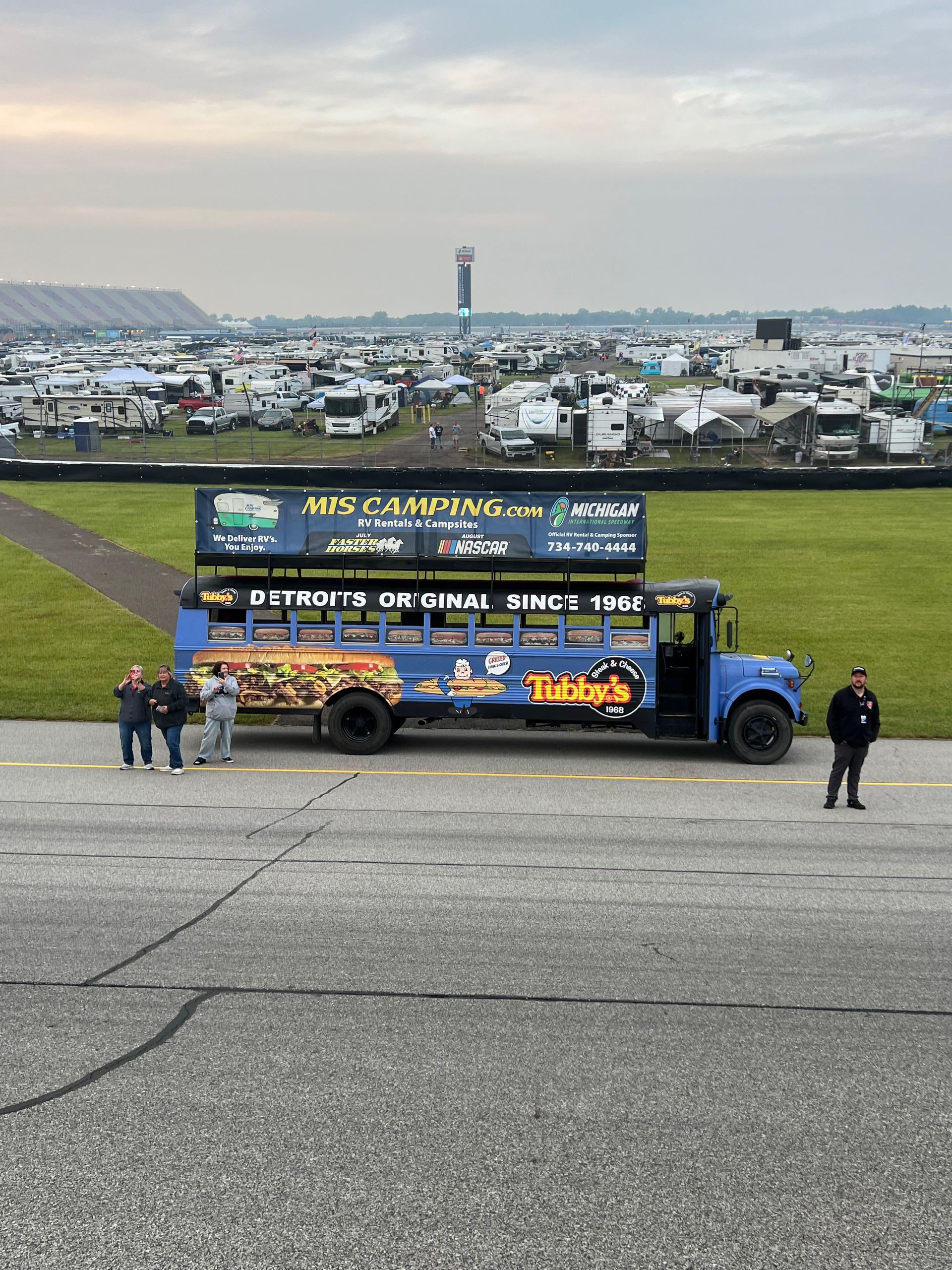 Blue bus with signage at racetrack with people standing around.