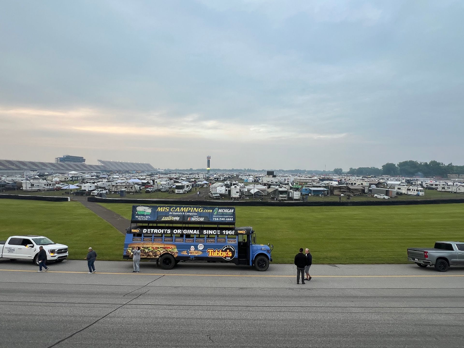 Large blue truck with advertisement at a racetrack with campers and vehicles under cloudy skies.