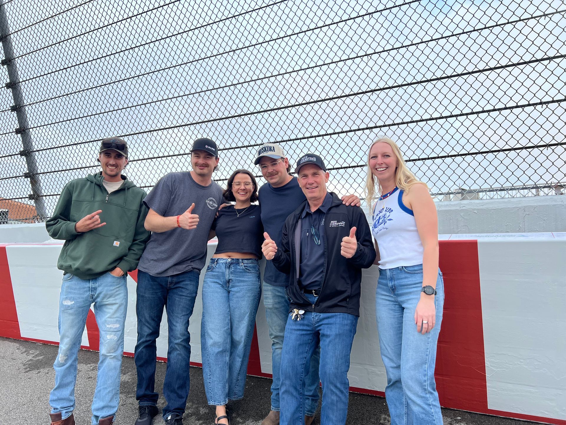 Group of six people smiling, giving thumbs up next to a red and white barrier and chain link fence.