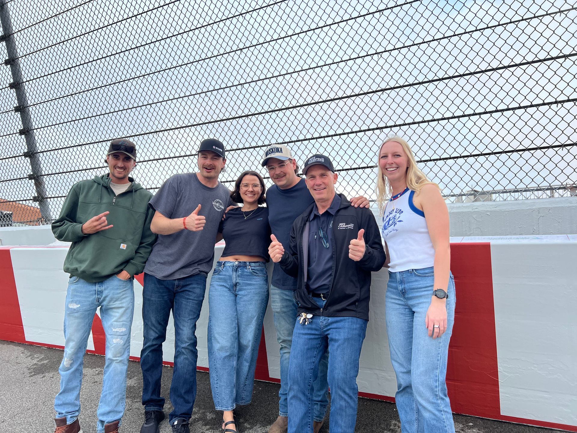 Six people smiling and giving thumbs up at a racetrack, standing by a wall with red and white stripes.