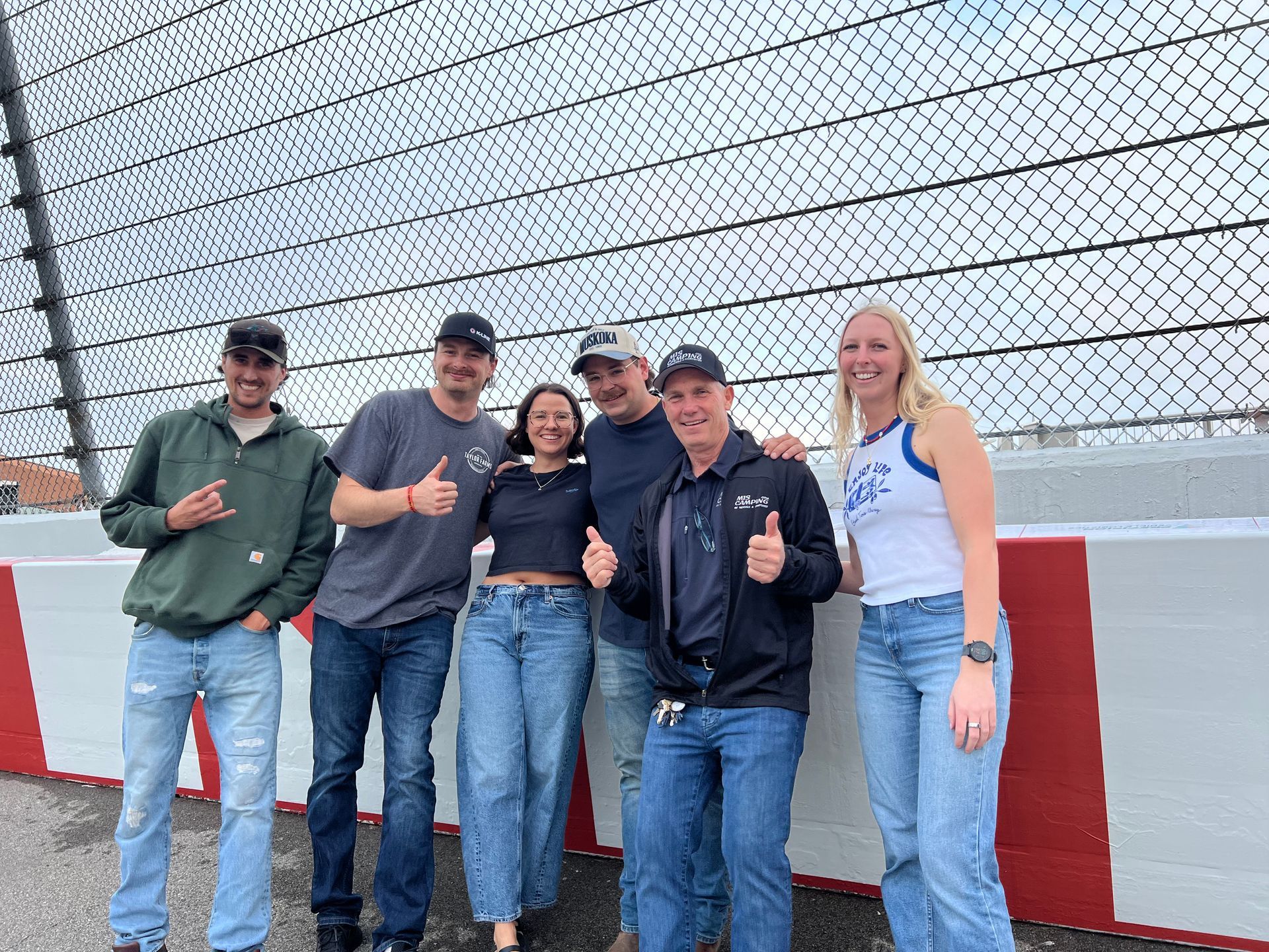 Six people smiling and giving thumbs up at a race track. They are standing near a wall, fence, and the sky.