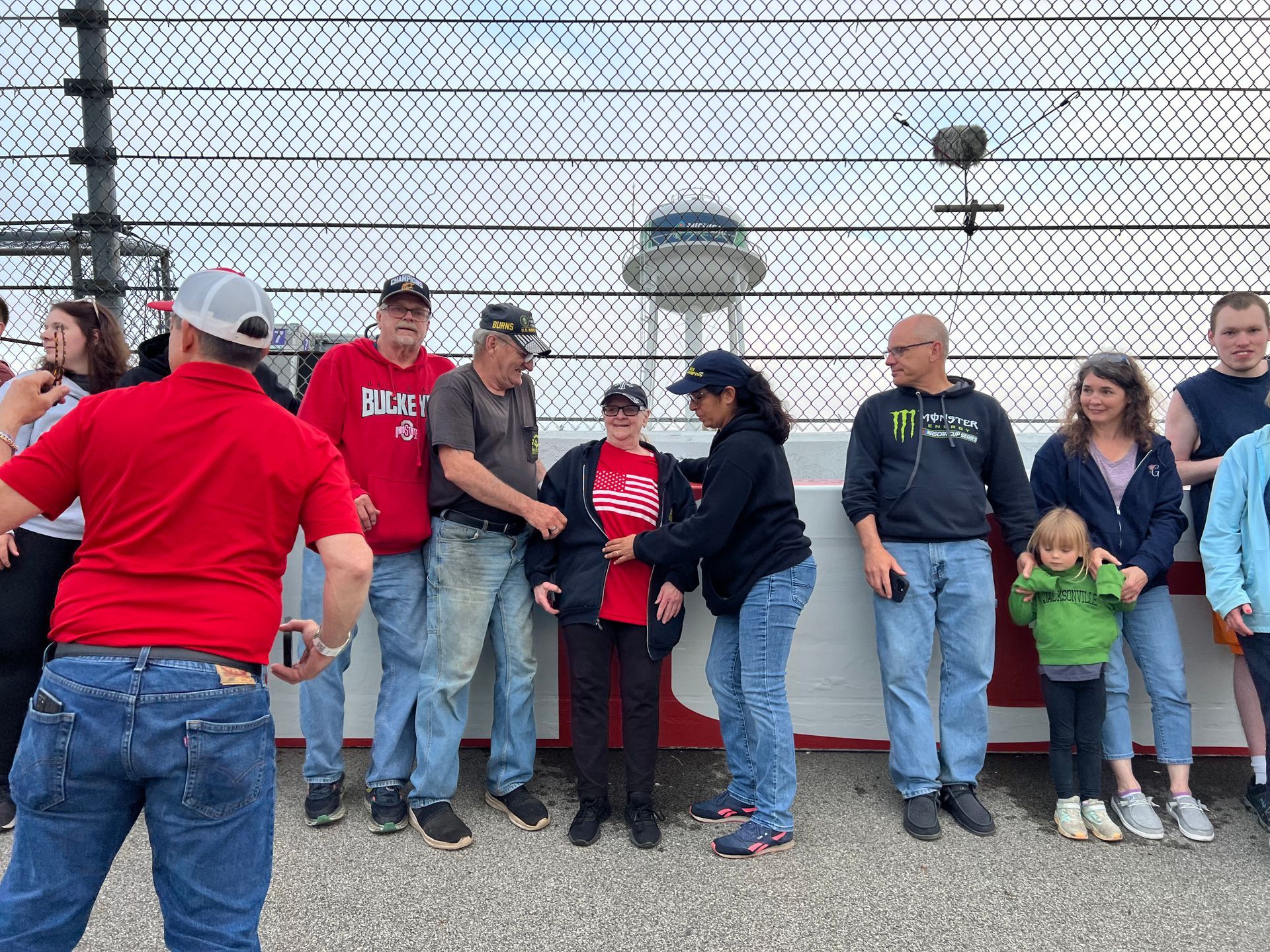 People at a racetrack stand near a fence; some shake hands.
