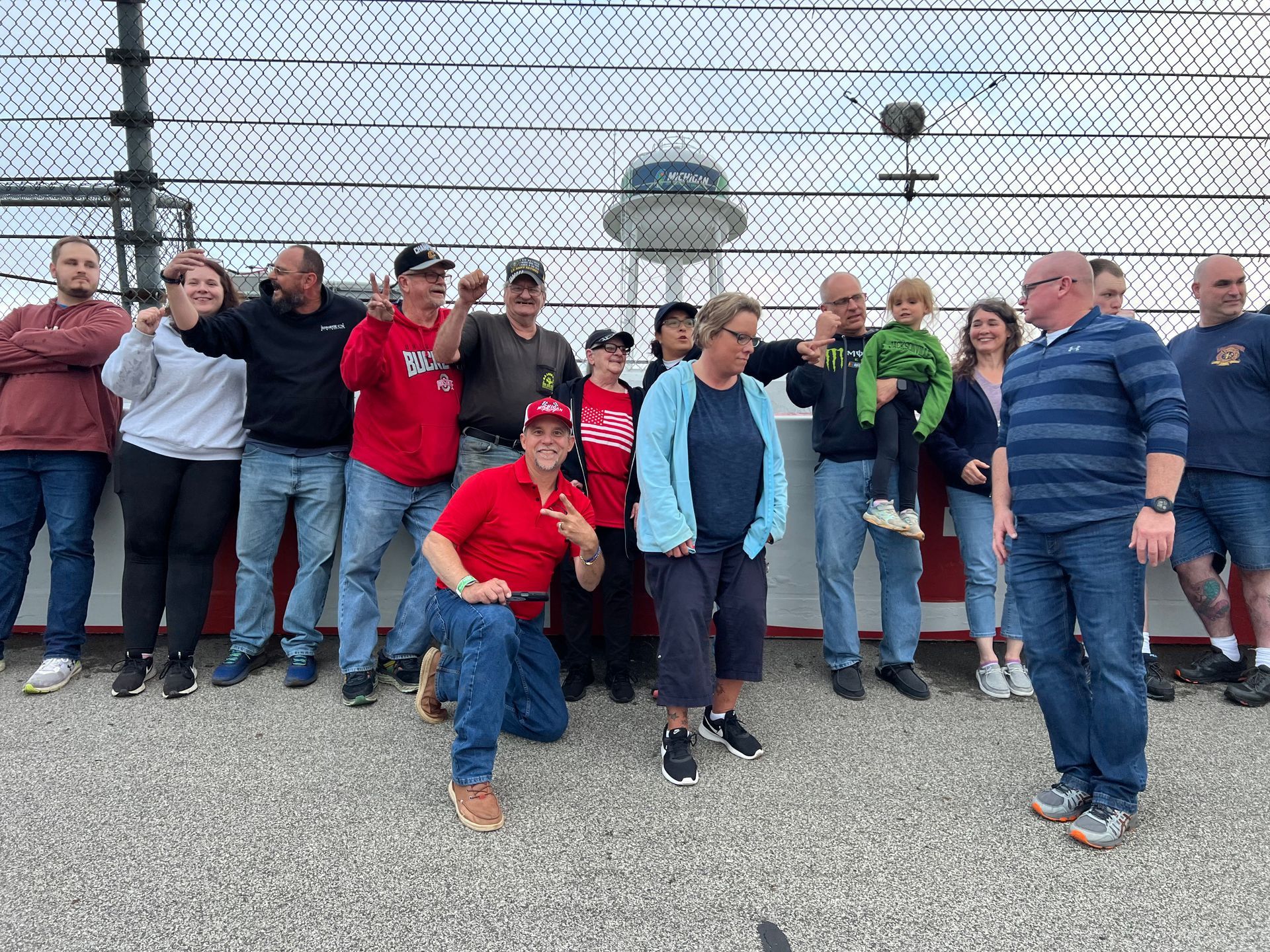 Group of people pose in front of a white and gray wall, some smiling and gesturing. Tower in the background.