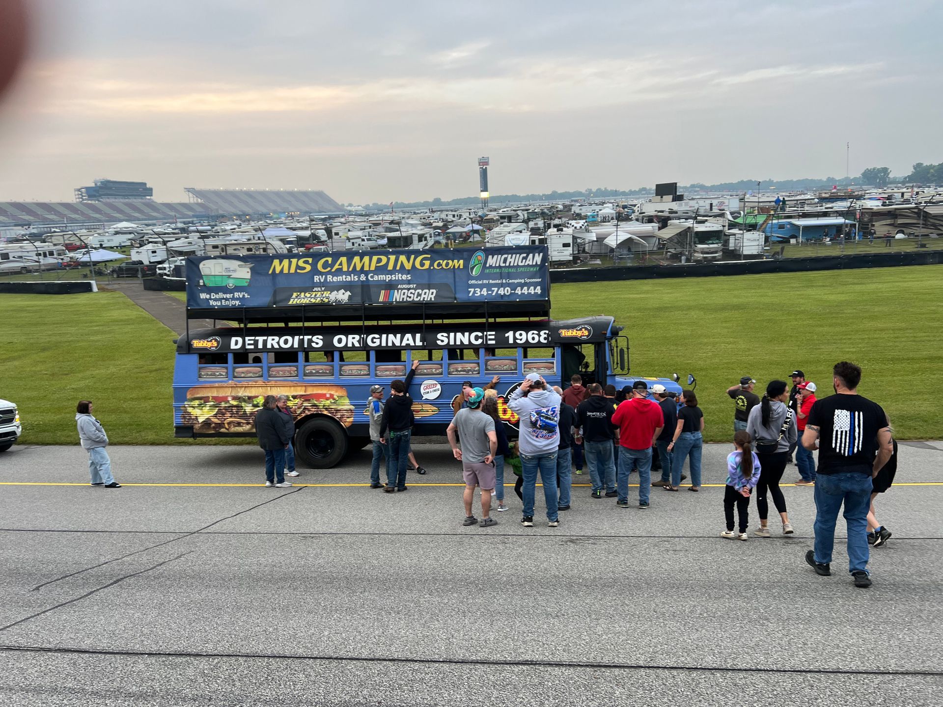 People gathered around a blue bus with signage; on a paved area, with campsites and track in the background.