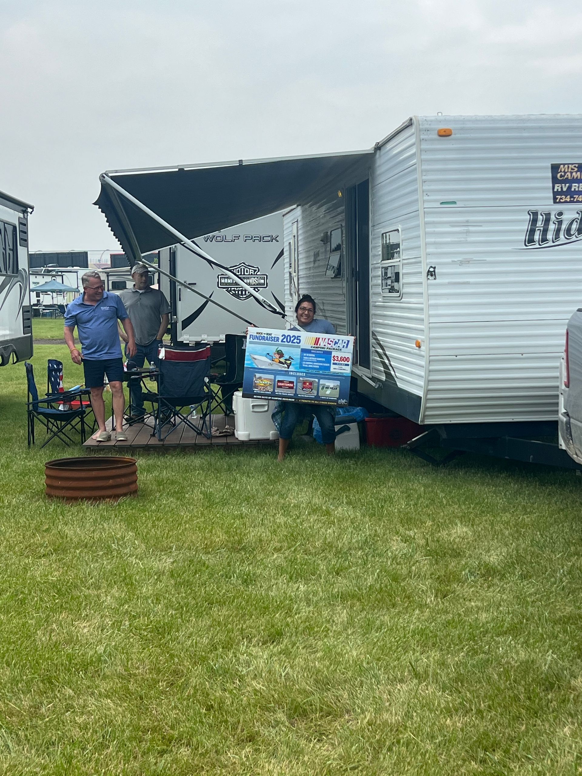 People outside a camper, one holding a box of items; green grass, awning extended.