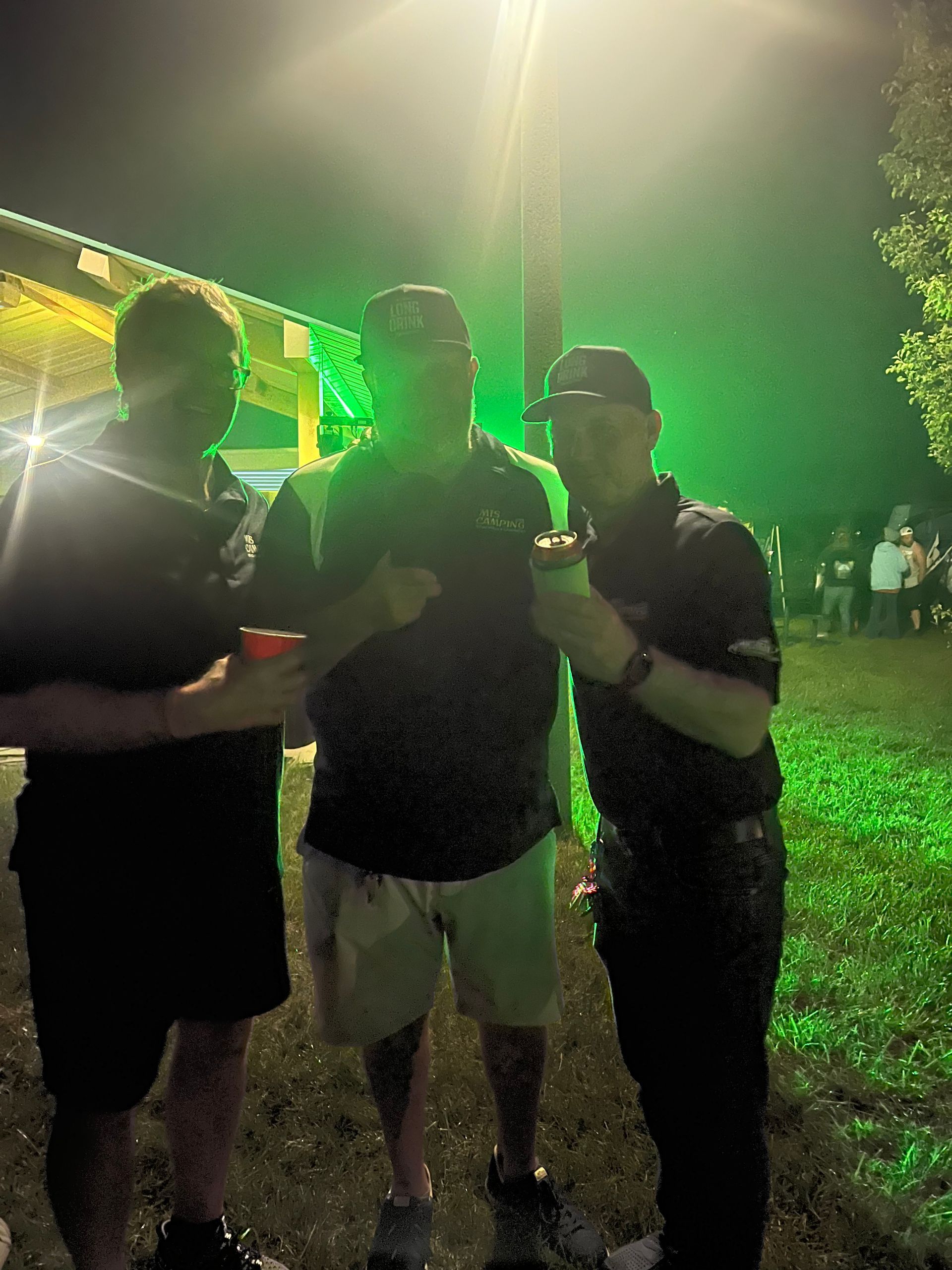 Three men holding drinks, posing outside at night under green light.