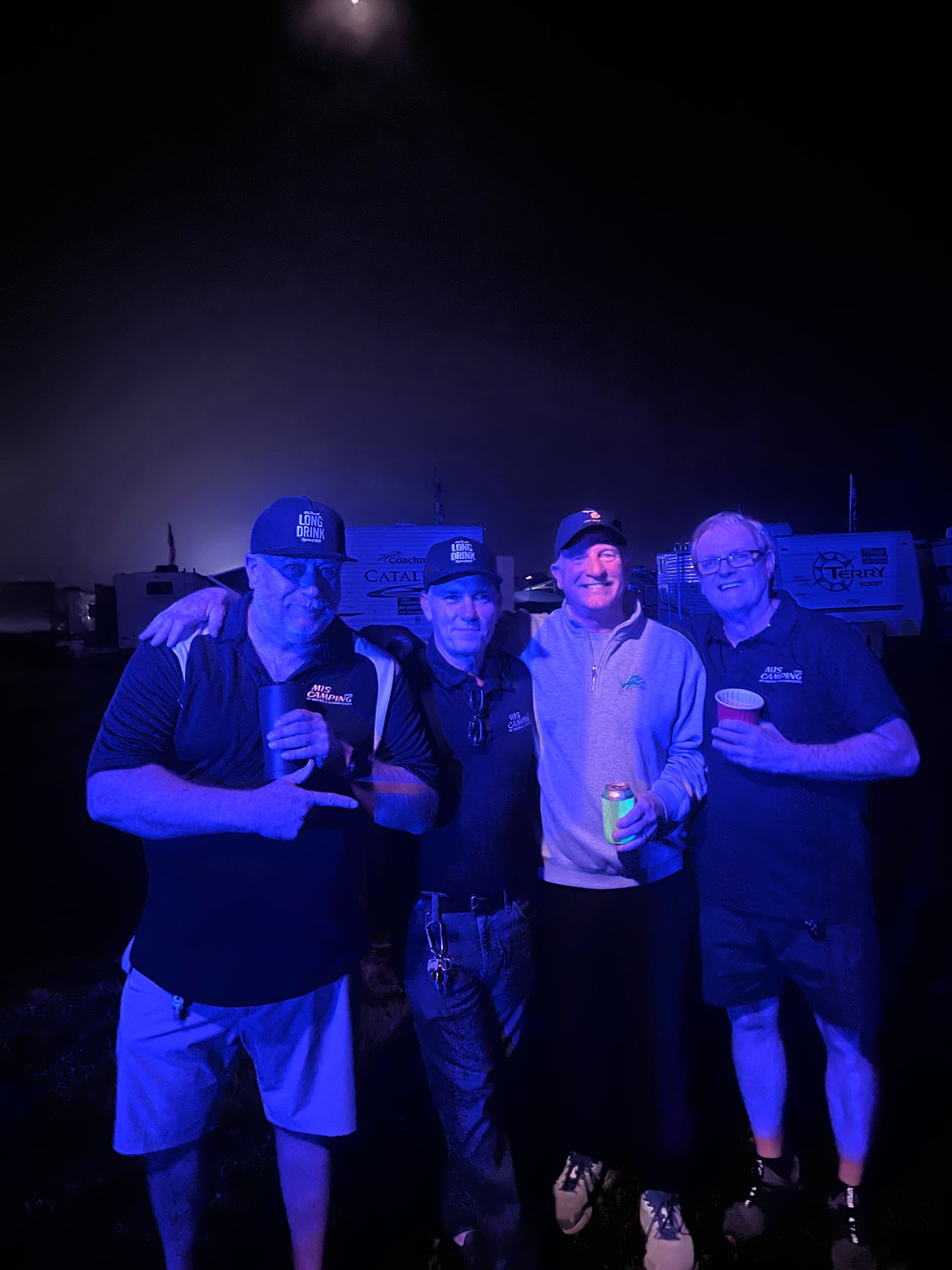 Four men in hats stand together at night, illuminated by blue light, possibly holding drinks.