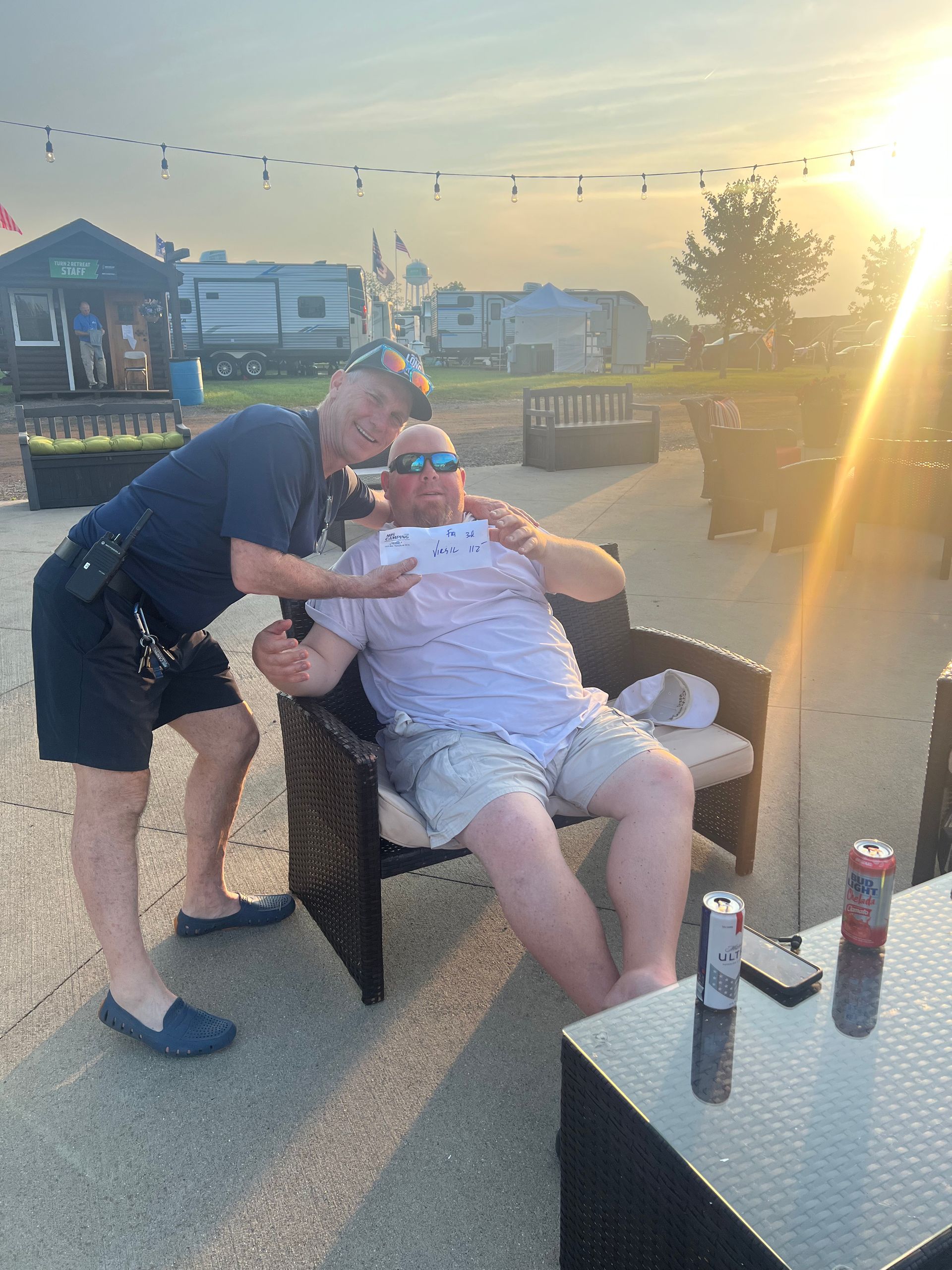Two men outdoors; one stands, points at the other seated in wicker chair. Sunset in background.