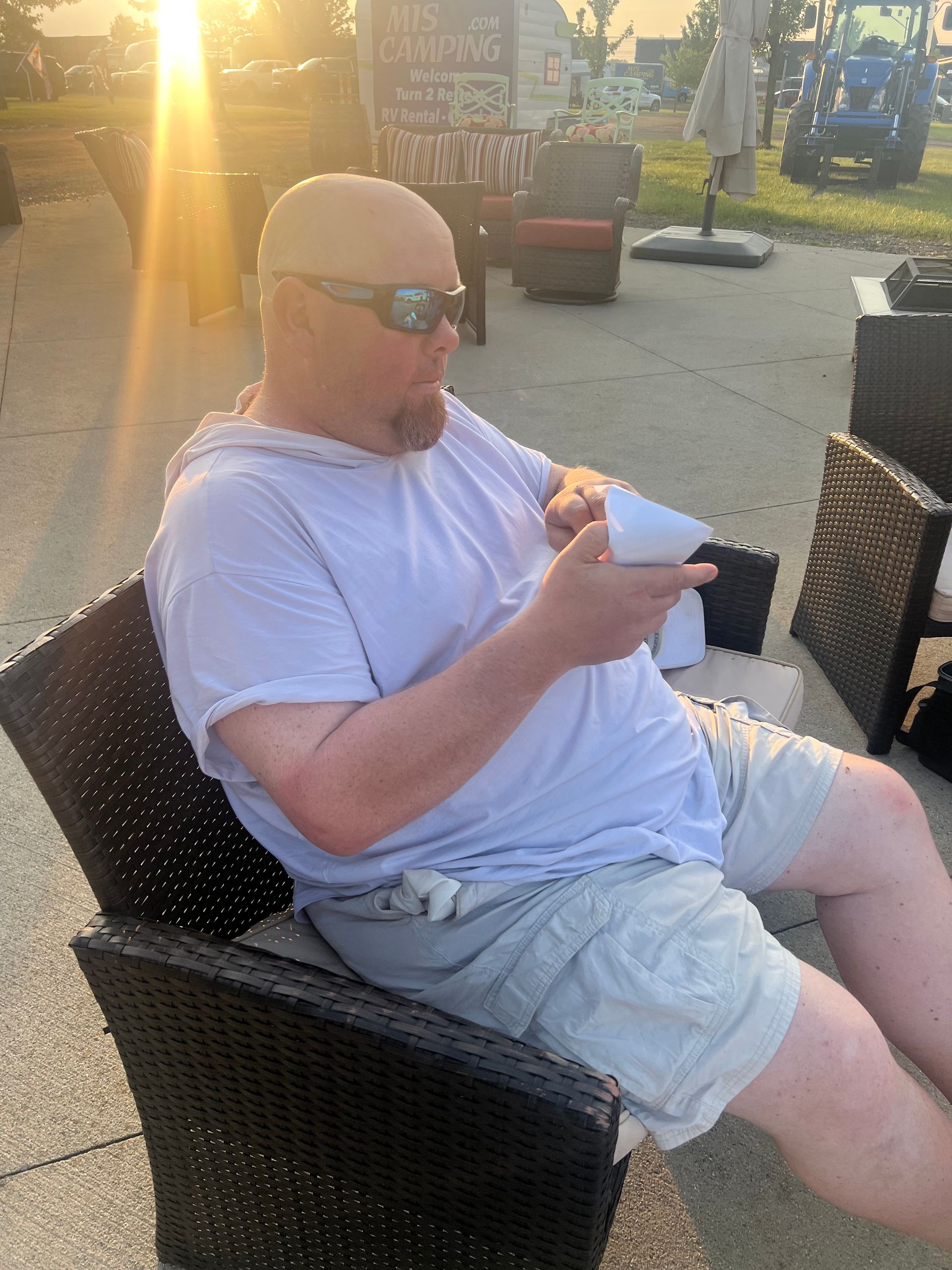 Man in sunglasses seated outdoors, holding paper; sunny background.