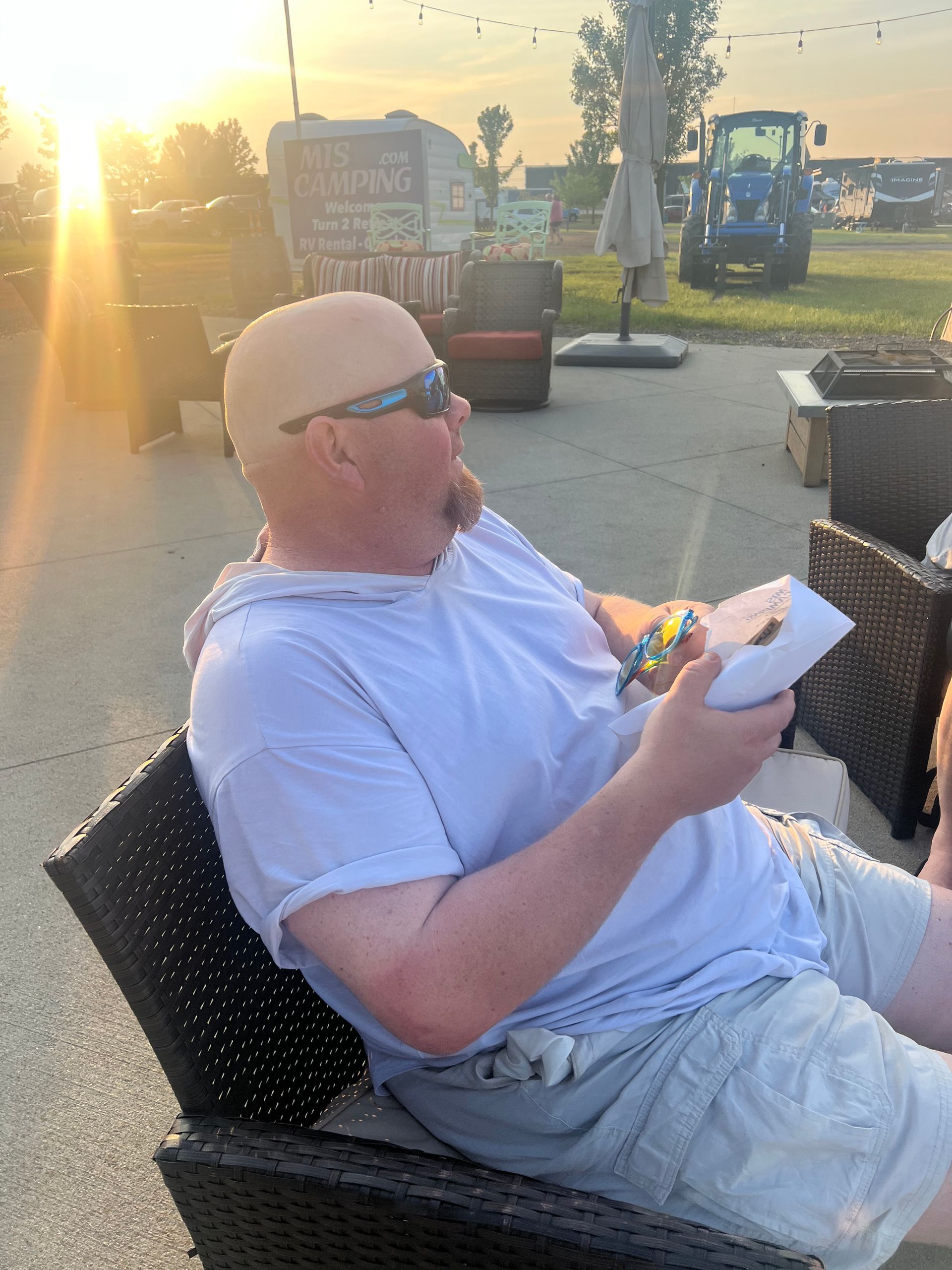 Man seated outdoors, holding food, looking up with sunset in background.