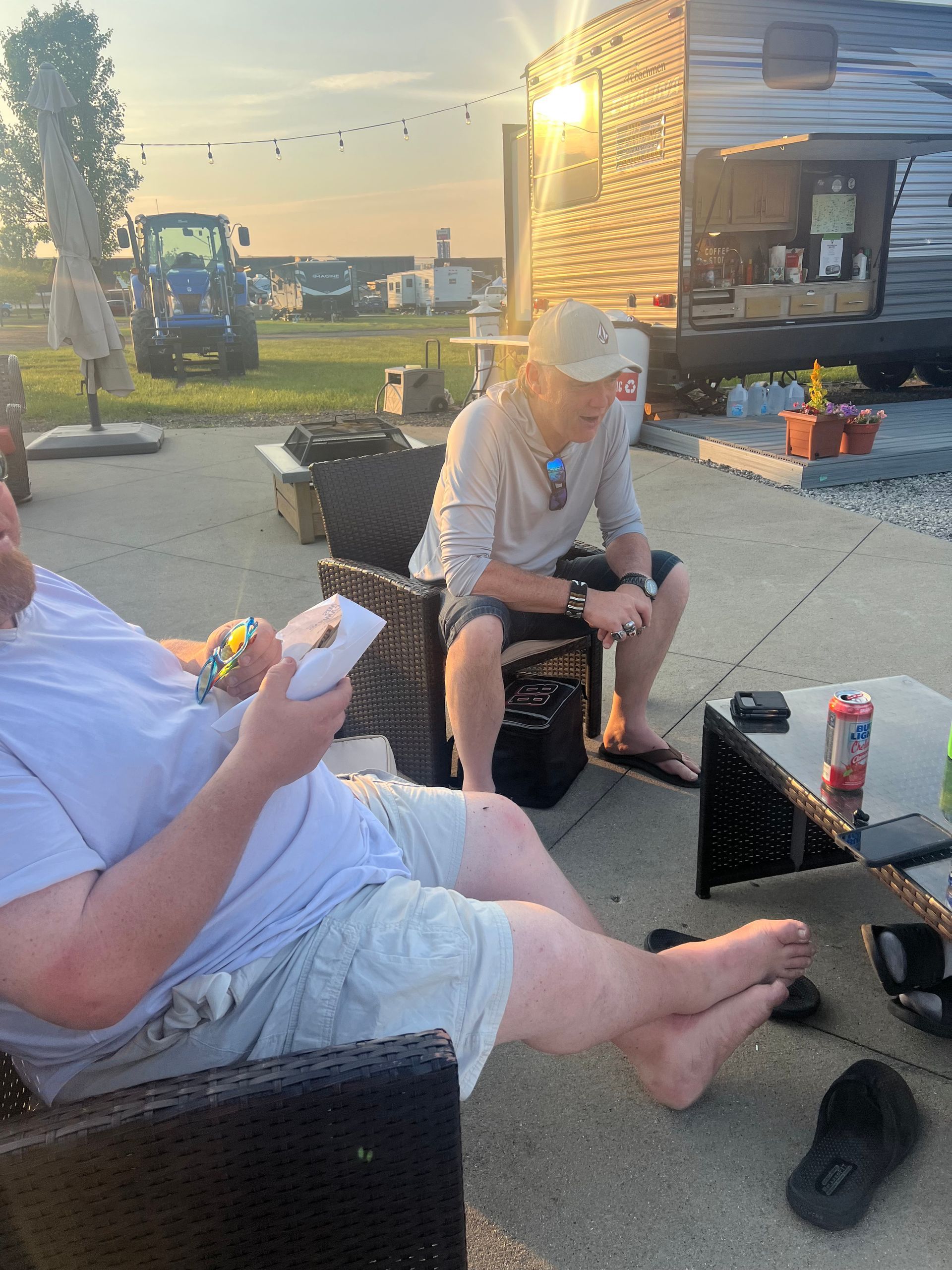 Two men sitting outside near a camper. One holds a plate; the other wears a cap. Sunset casts warm light.