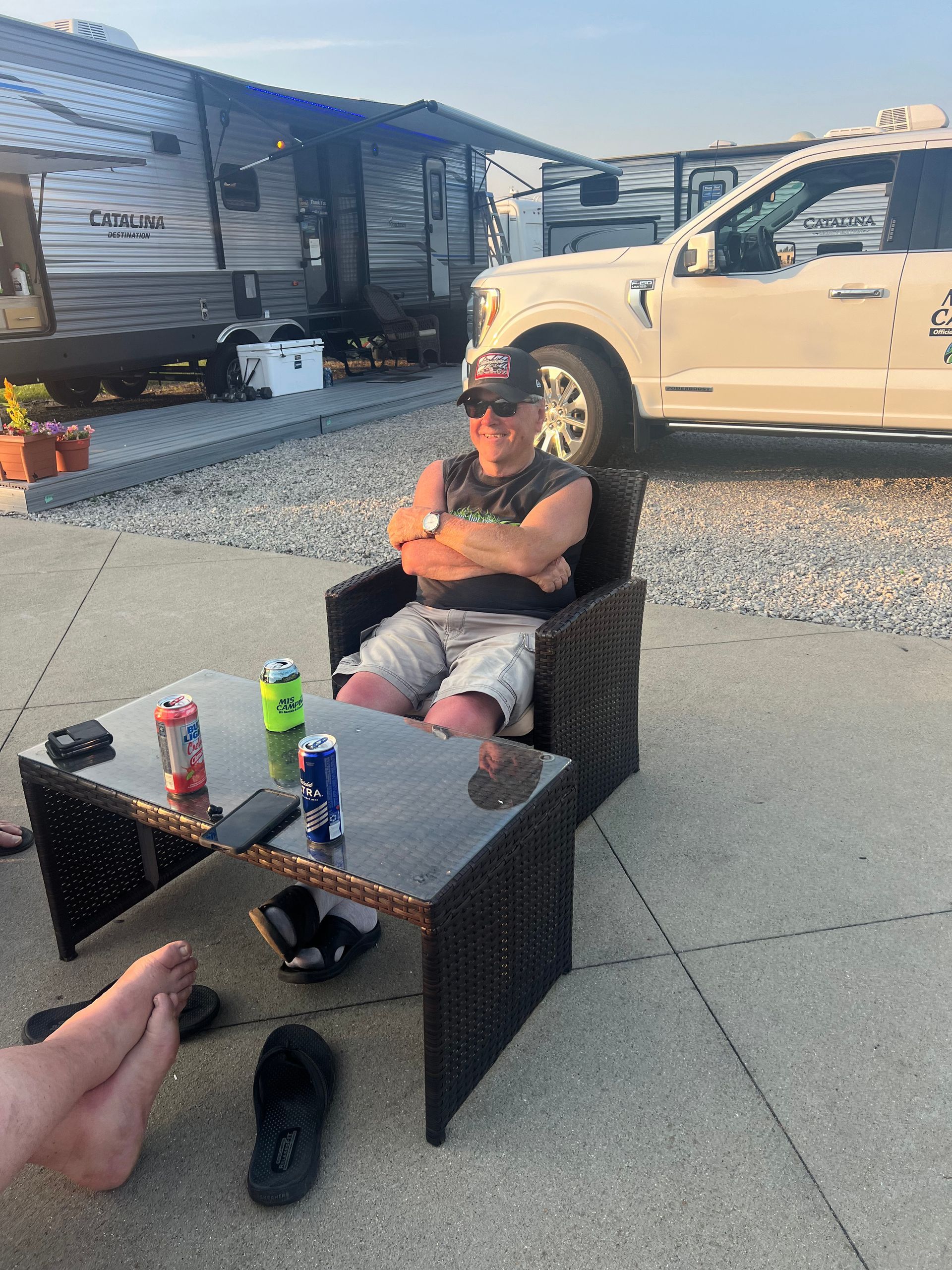 Man sitting in chair, arms crossed, at a campsite with drinks on table.