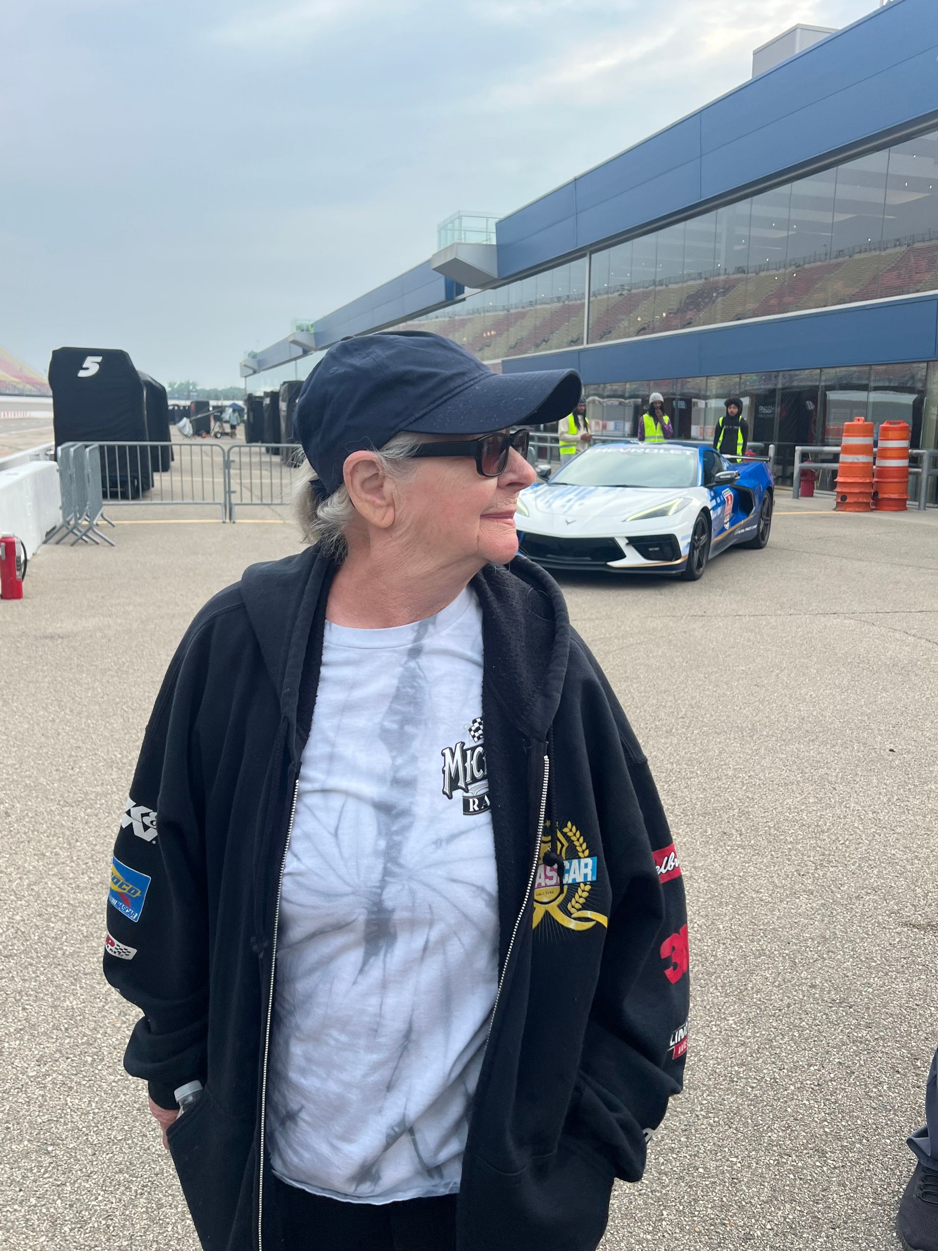 Woman wearing a hat and sunglasses, standing near a racetrack with a white sports car.