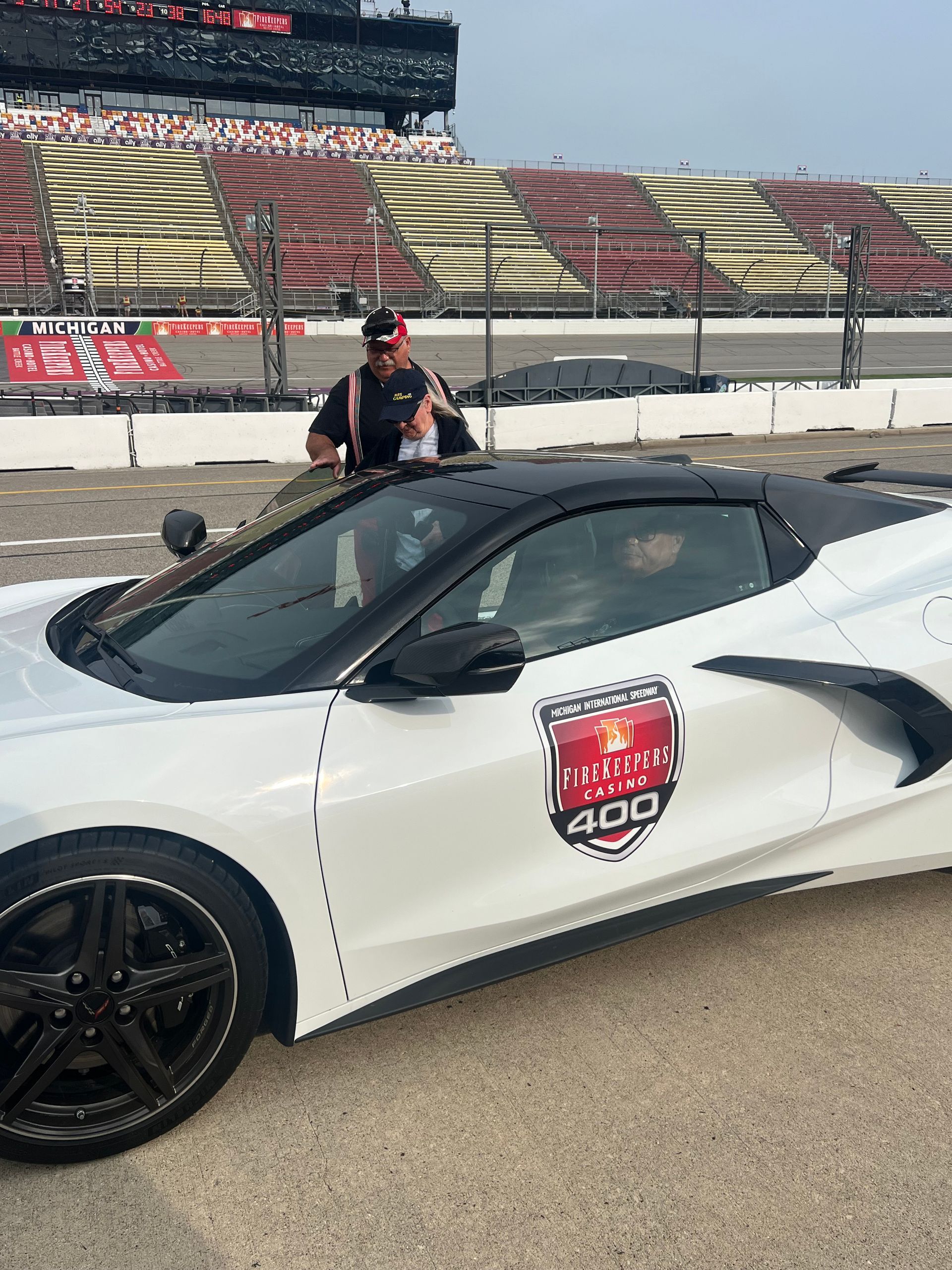 White Corvette on racetrack with person holding trophy and grandstand in the background.