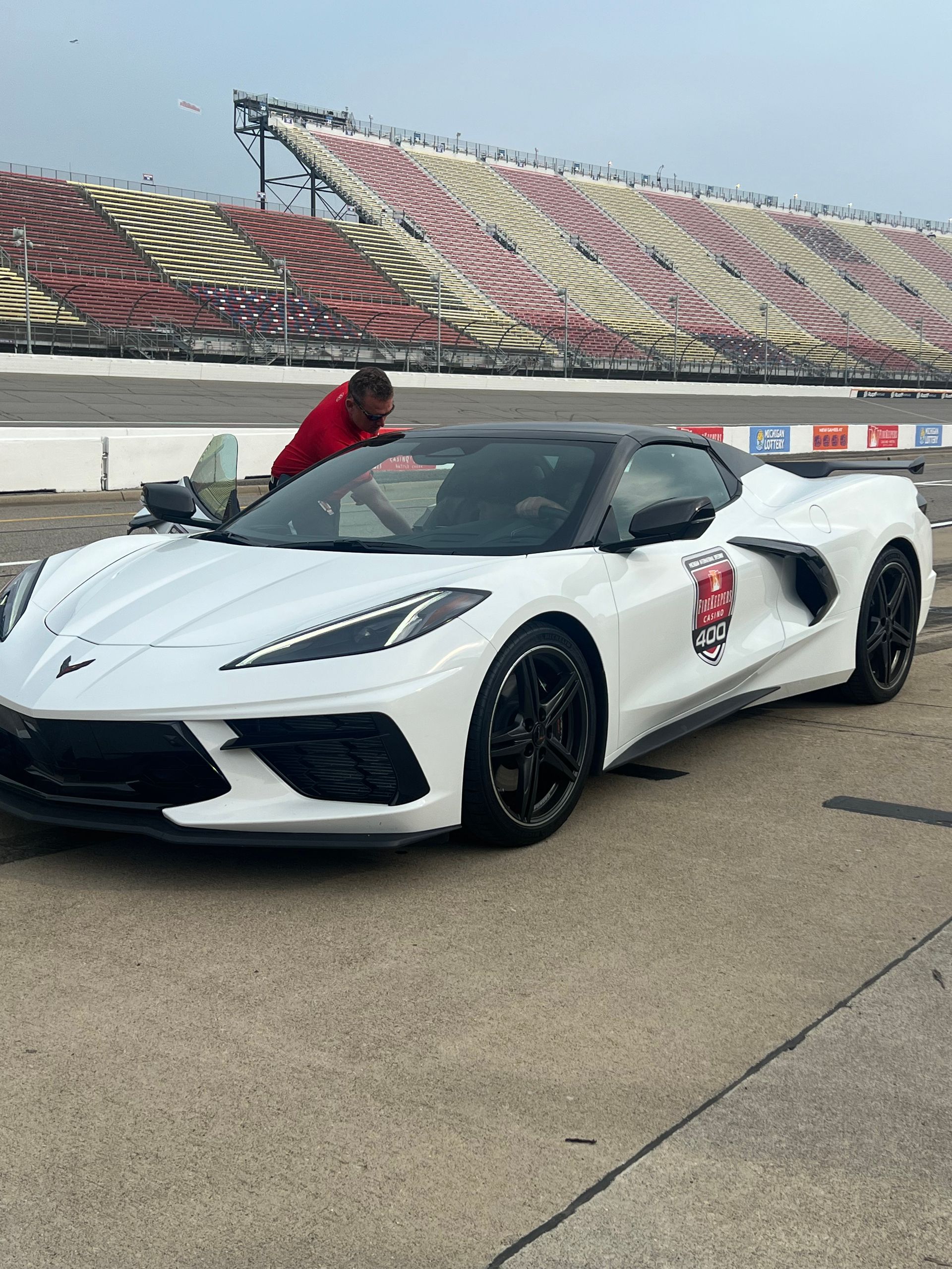 White Corvette convertible on a racetrack, person by the driver's side door, grandstand in the background.