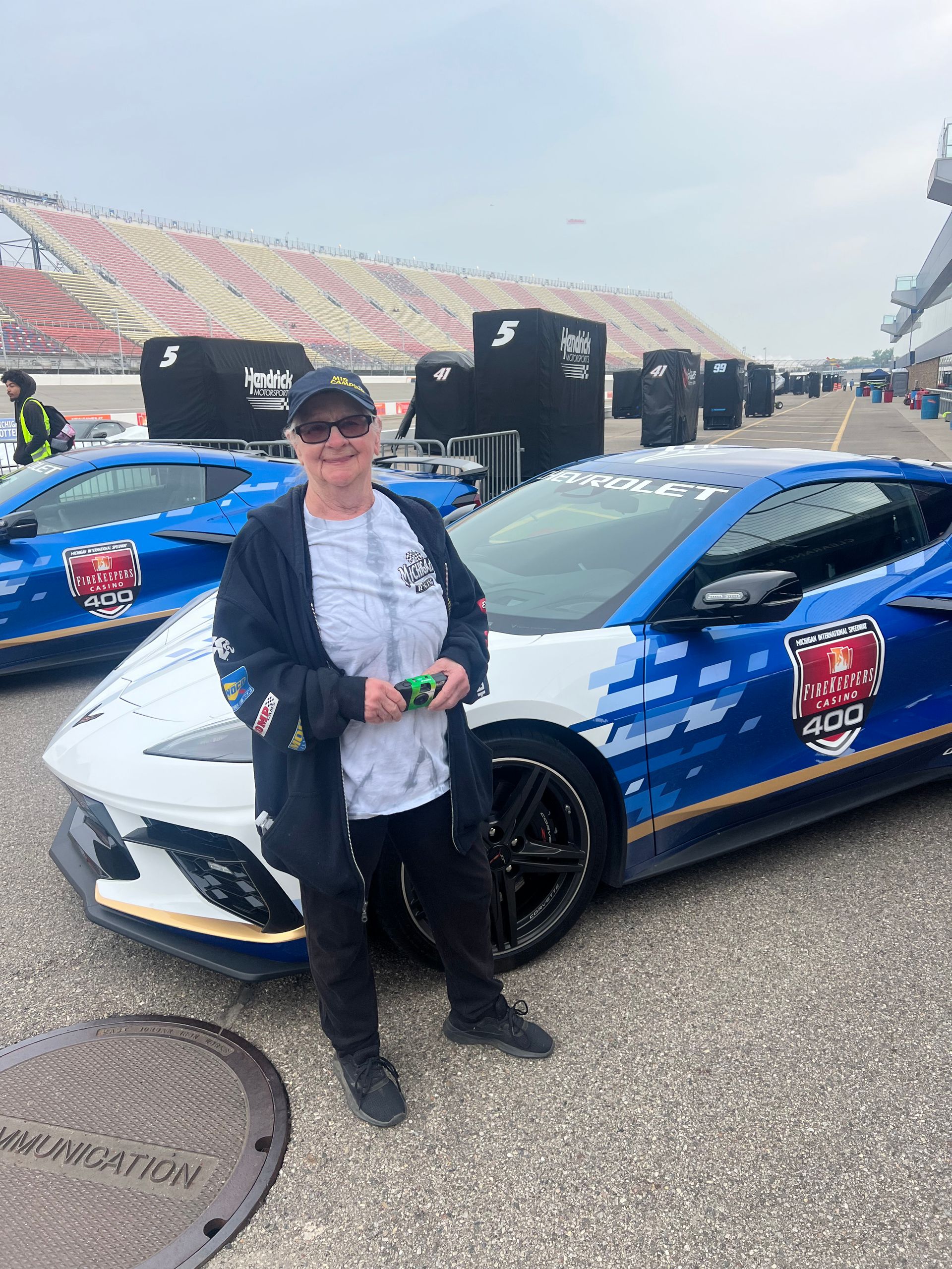 Person stands next to a blue and white sports car at a racetrack, wearing a jacket and hat.