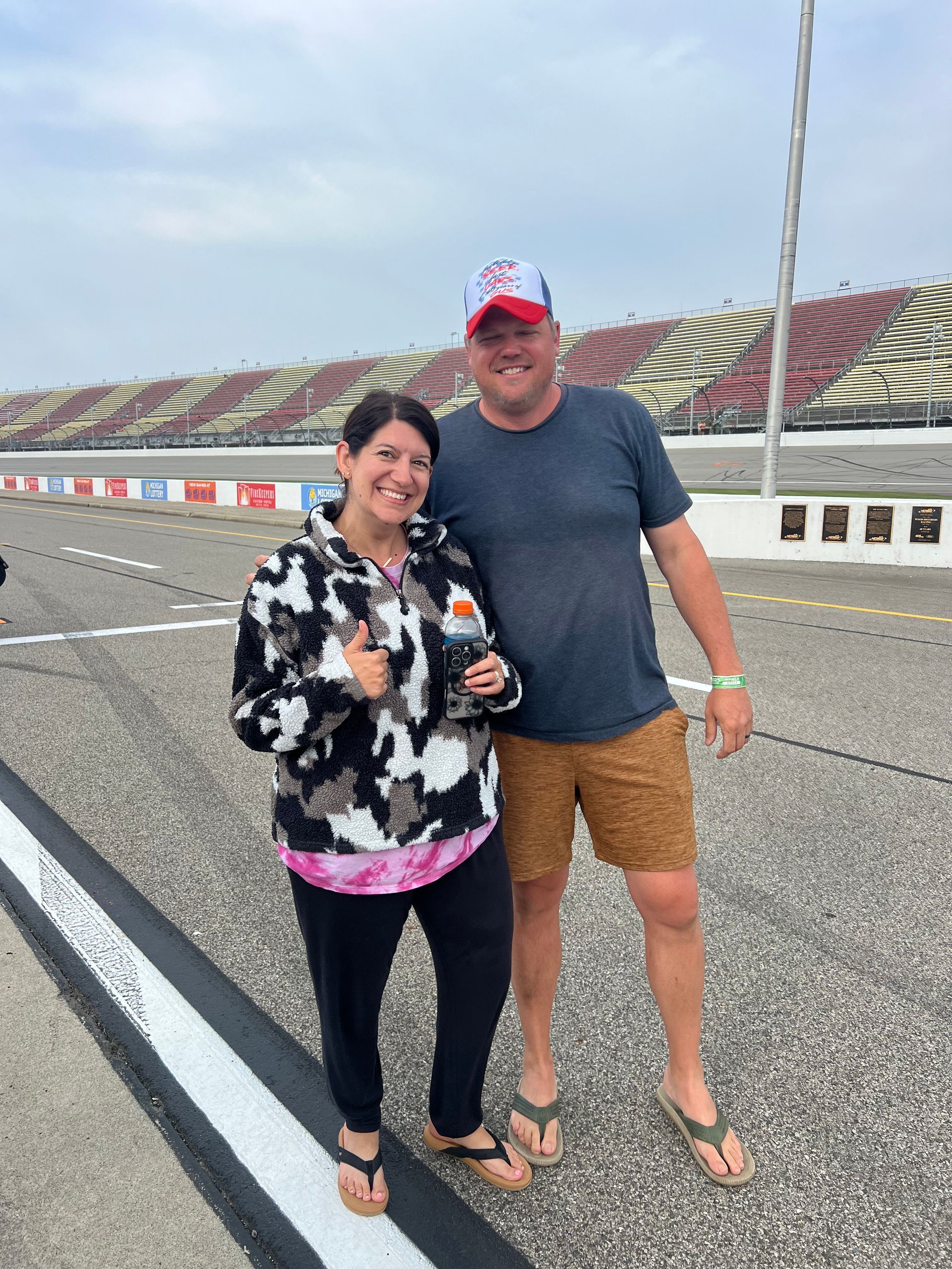 Two people pose near a racetrack. Woman in black and patterned jacket gives thumbs up. Man in blue shirt and shorts smiles.