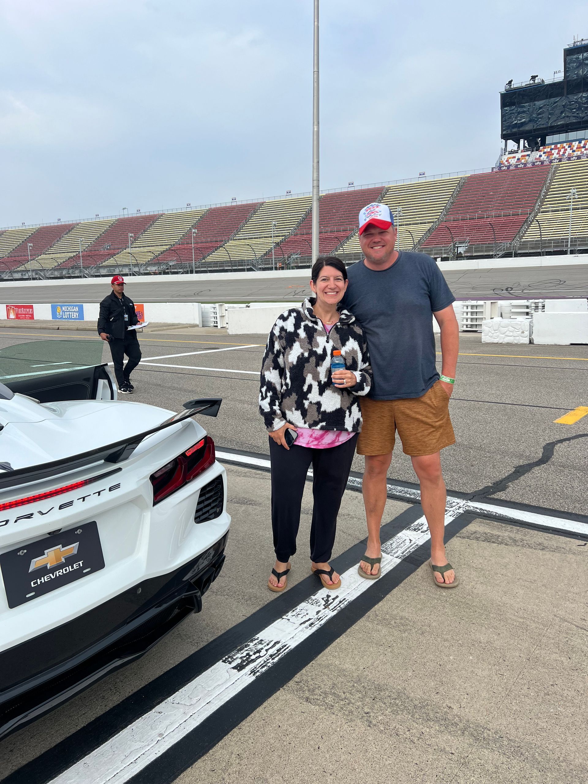 Couple poses next to a white sports car on a racetrack. Bleachers in the background.