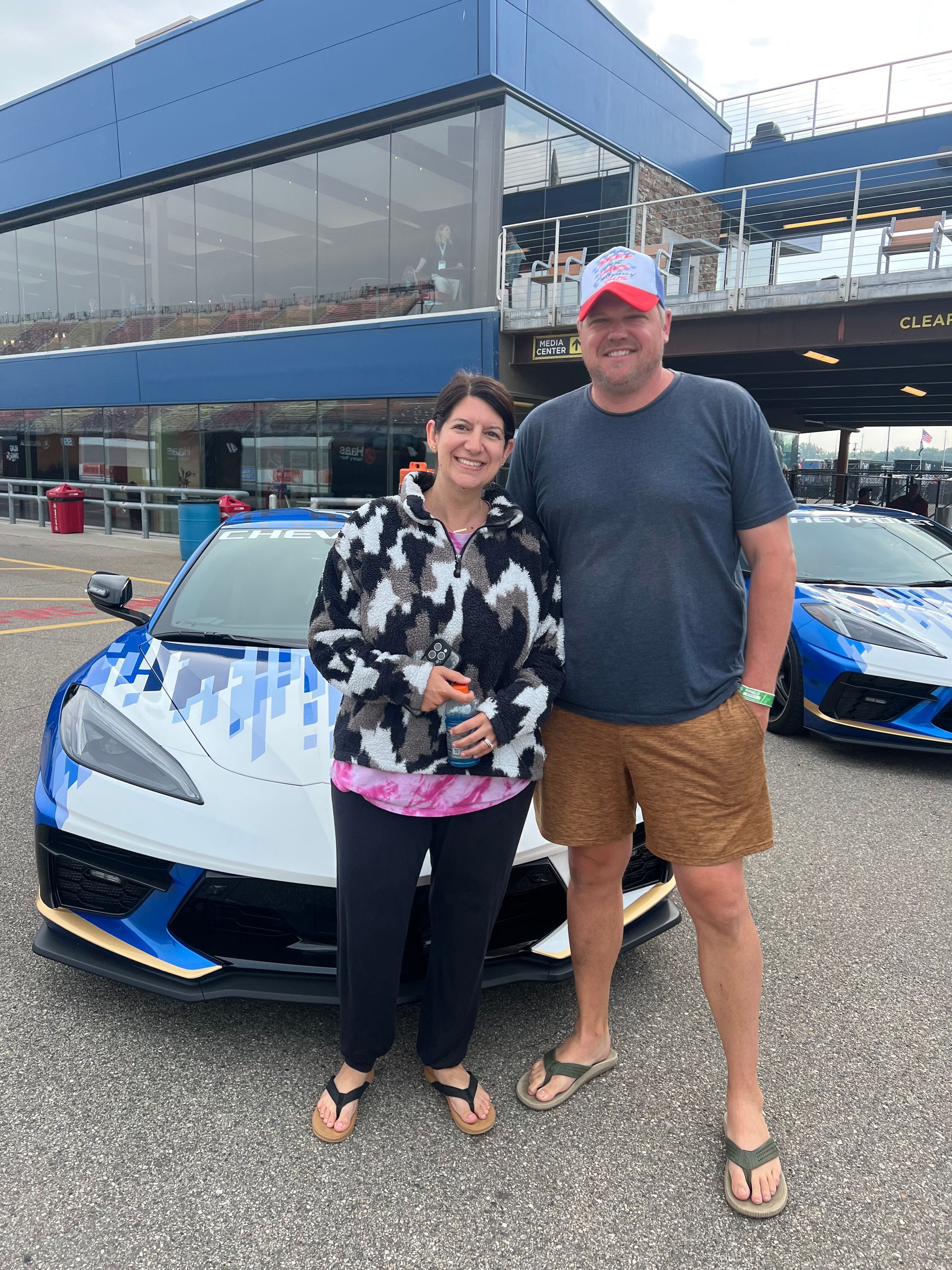 Man and woman standing by a blue and white sports car at a racetrack, smiles, sunny day.