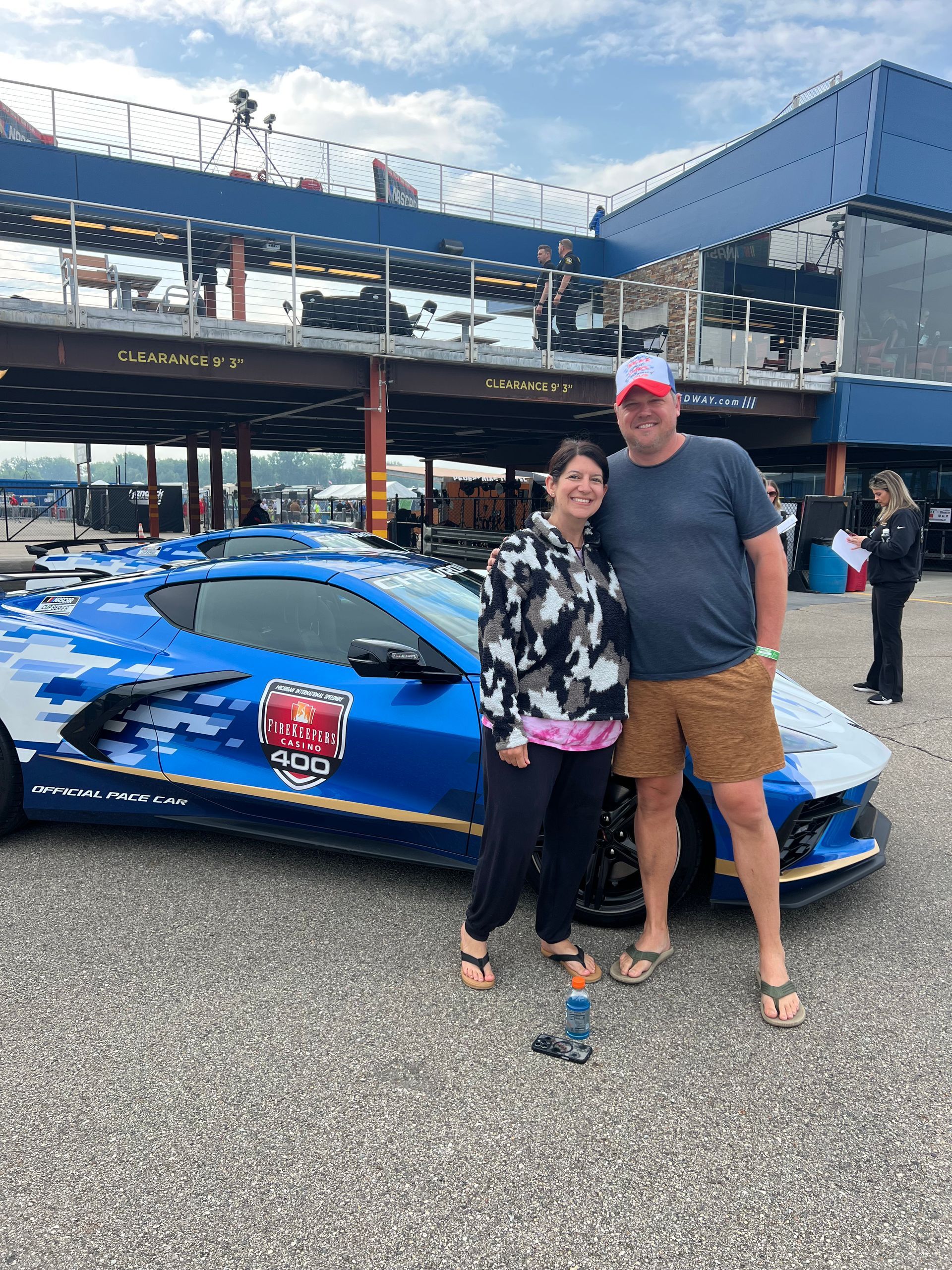 Couple posing with a blue camouflaged sports car at a racetrack, sunny day.