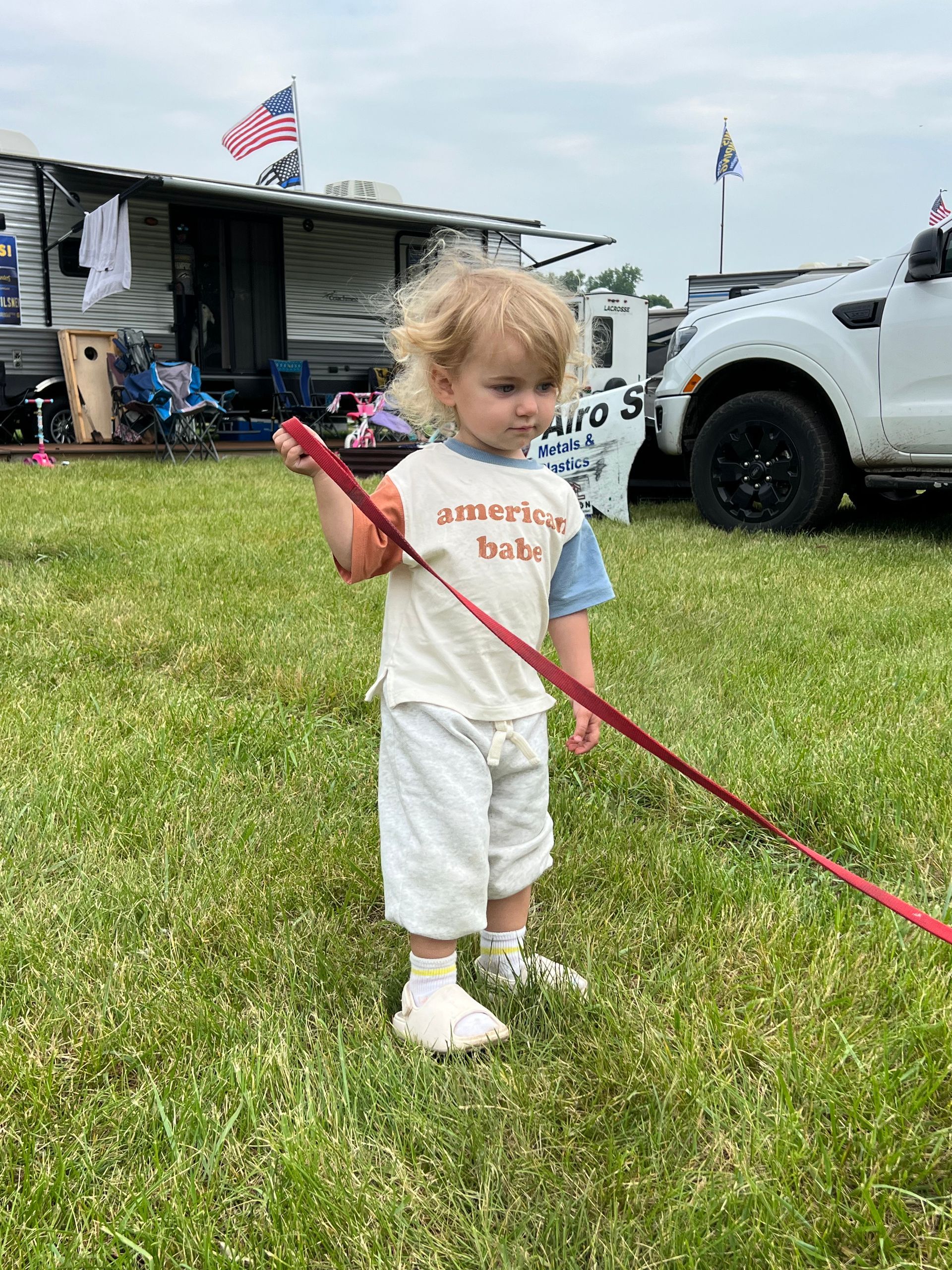 Young child holding a red leash, standing in grass near a trailer and a truck.
