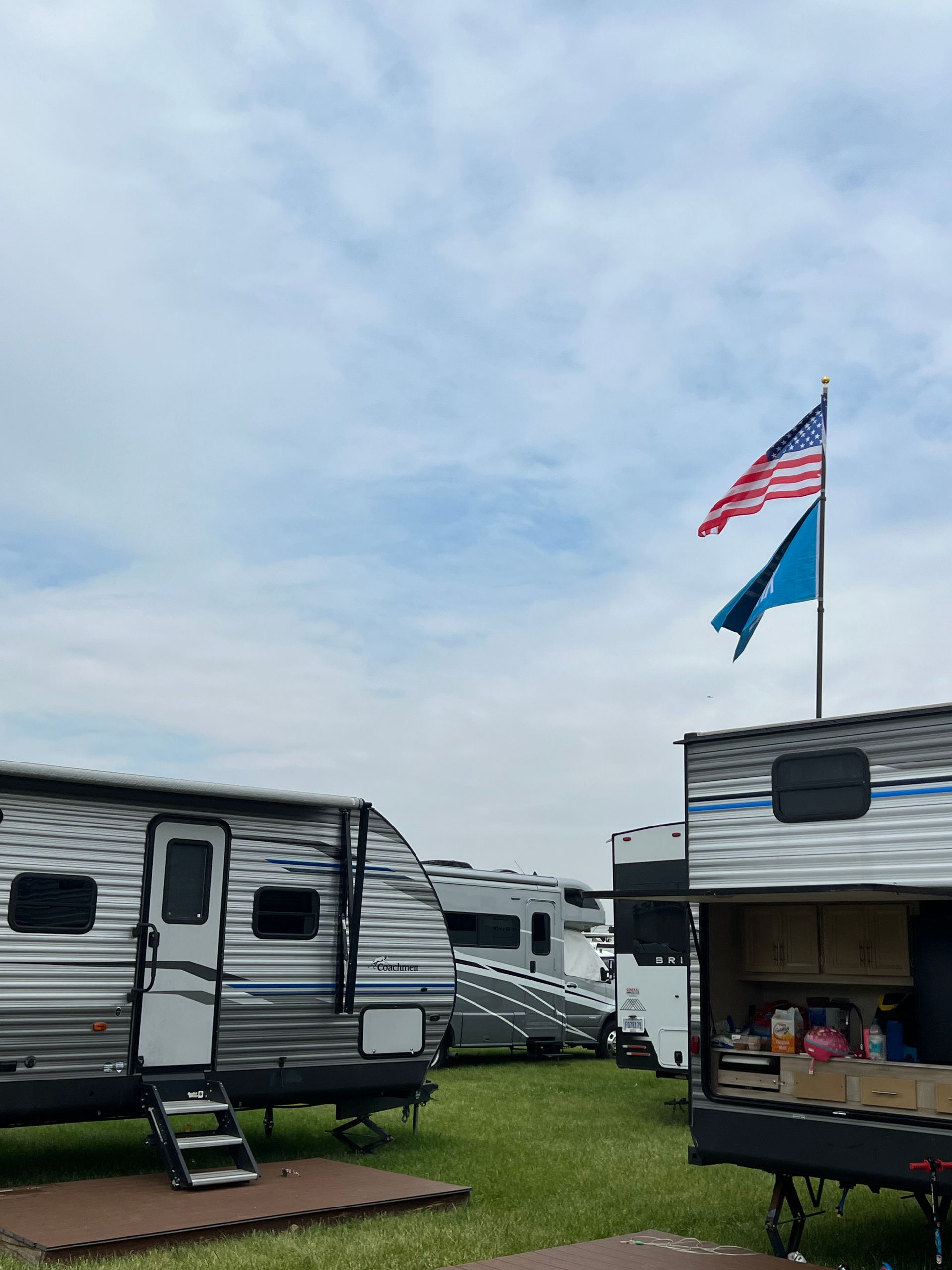Campground with RVs under a cloudy sky; American and blue flags fly.