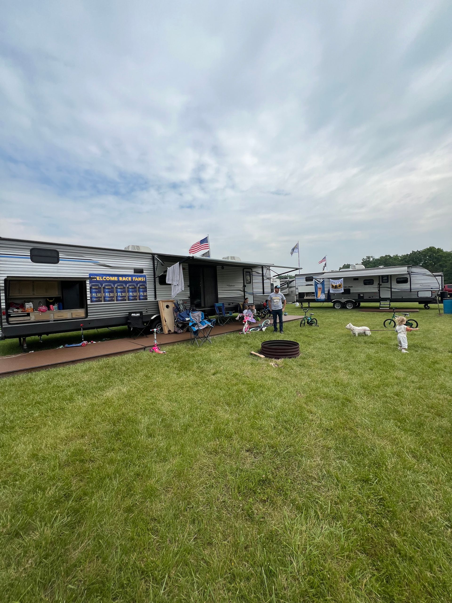 Campground scene with several RVs parked on green grass under a cloudy sky. People and small dogs are visible.