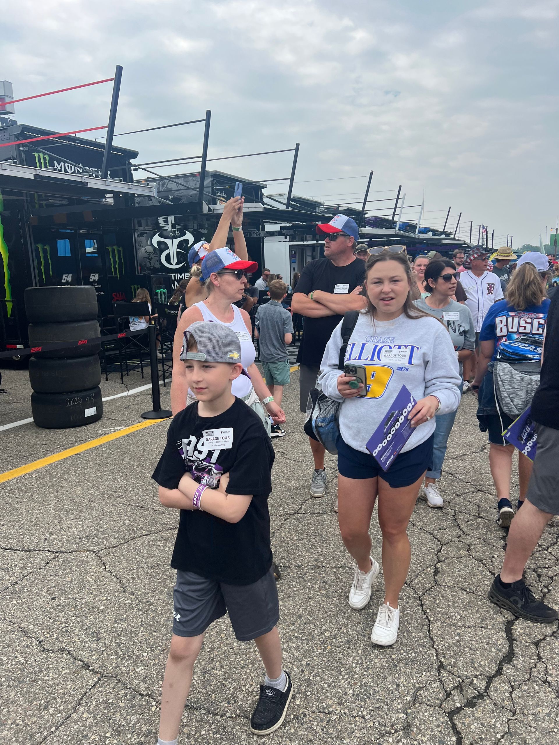 People walking near a race car garage. A boy in a cap and black shirt and a girl holding a cup and flag are in focus.