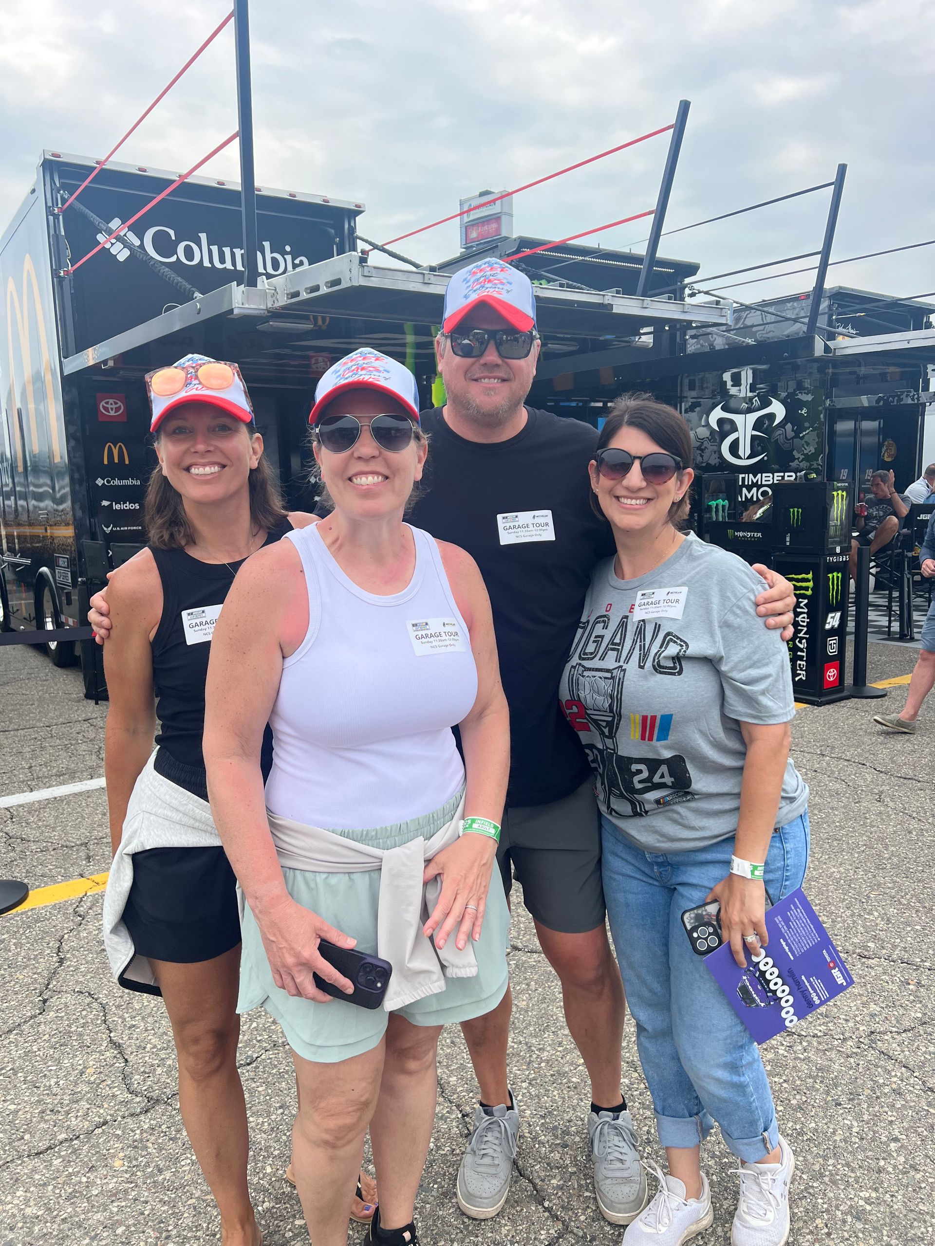Four people pose for a photo in front of a black trailer. They wear hats and sunglasses, smiling.