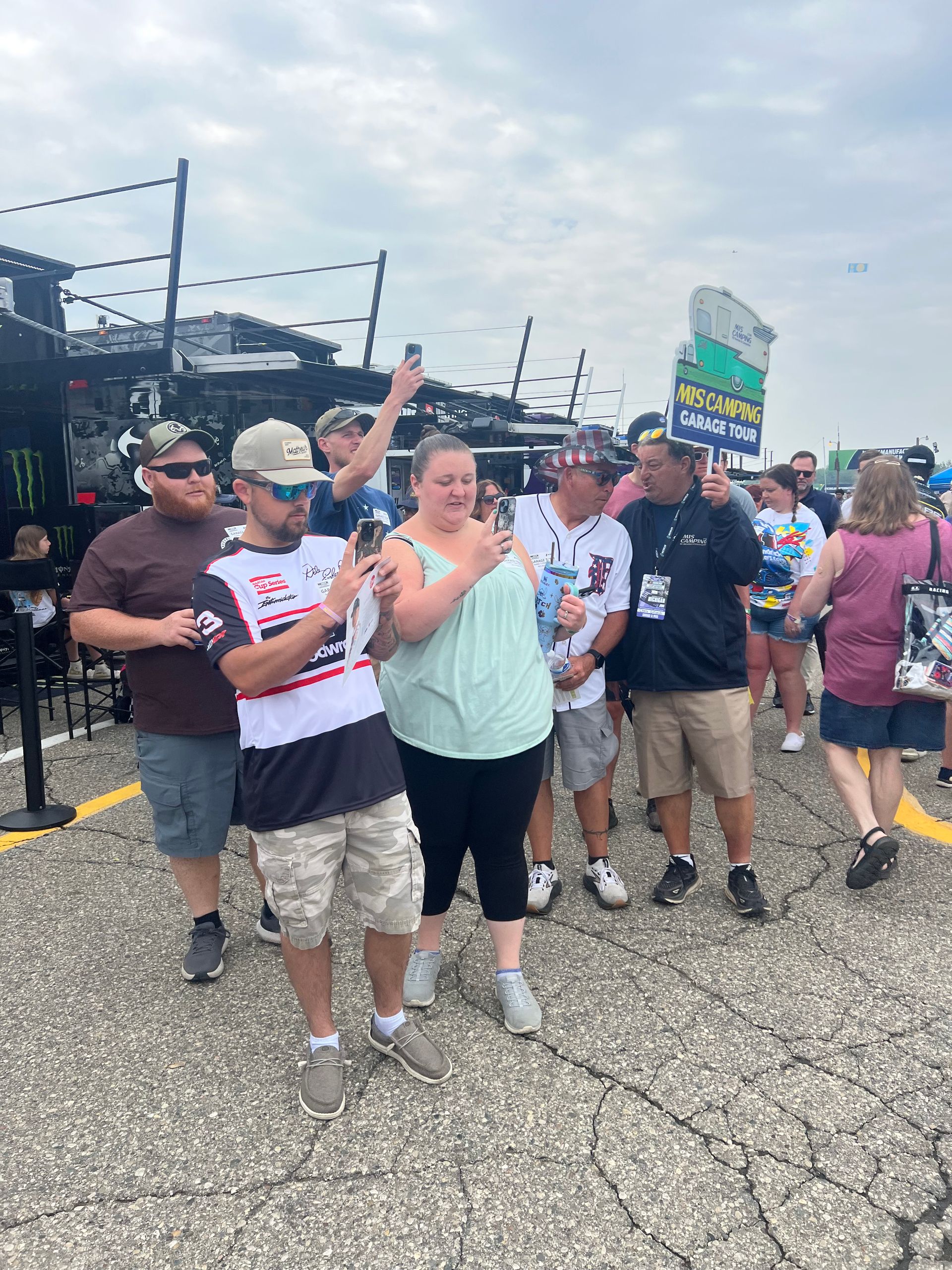Group of people near a black trailer, some holding phones and smiling, outdoors on a cloudy day.
