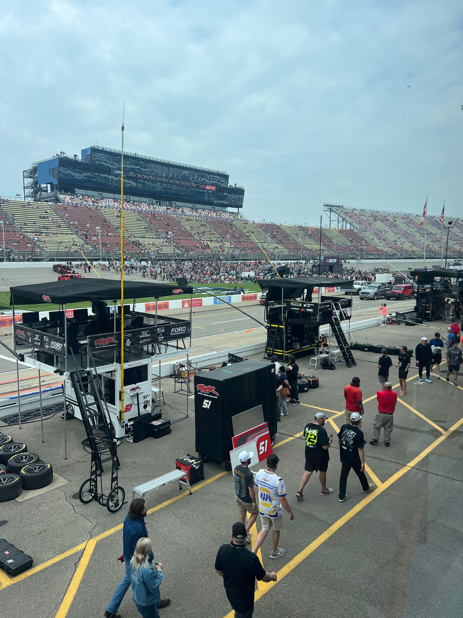 Race track scene with crew members, equipment, and a stadium full of spectators under a cloudy sky.