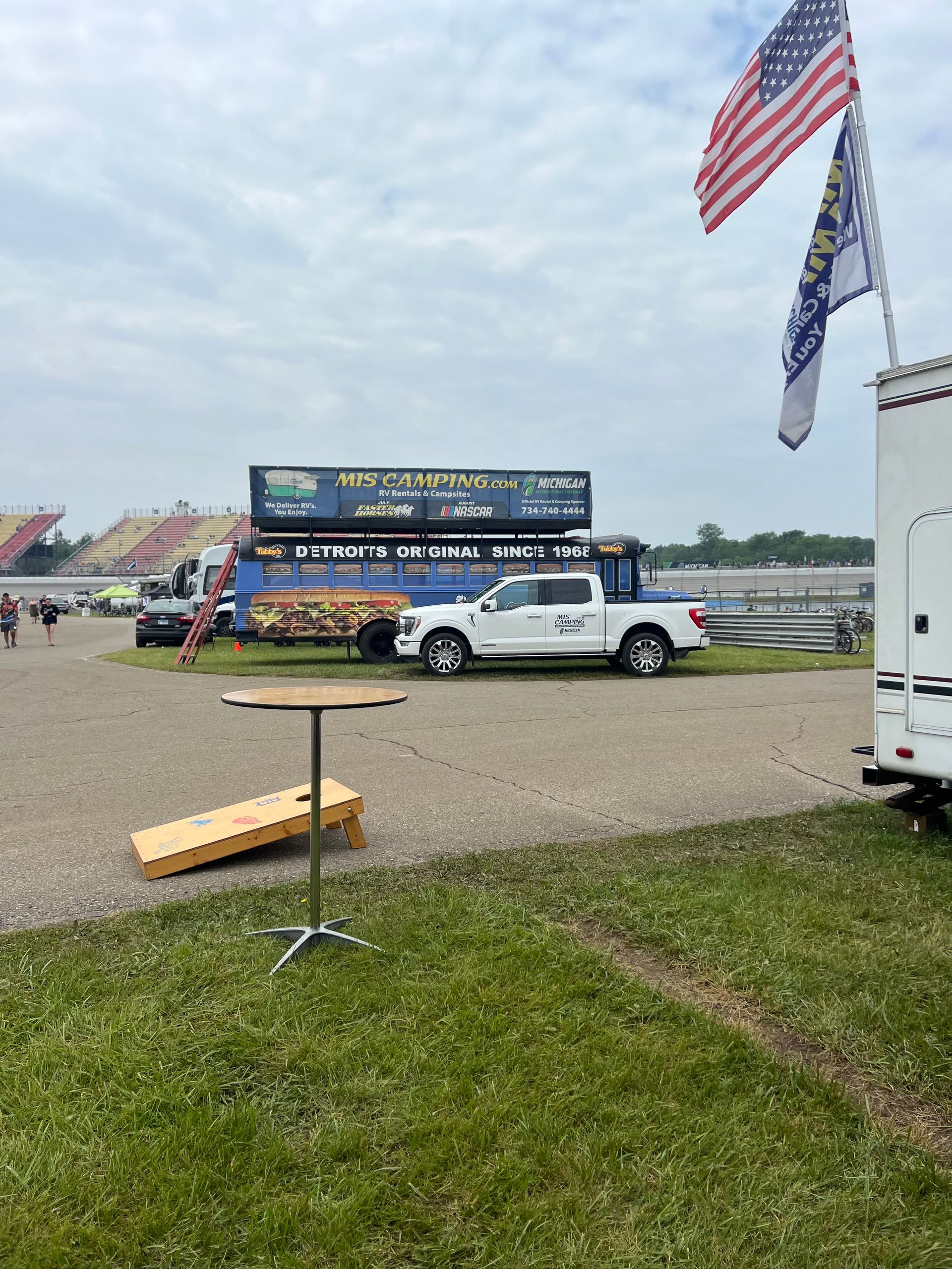 A white truck and a blue bus with American flags in front of a racetrack, with a cornhole set in the foreground.